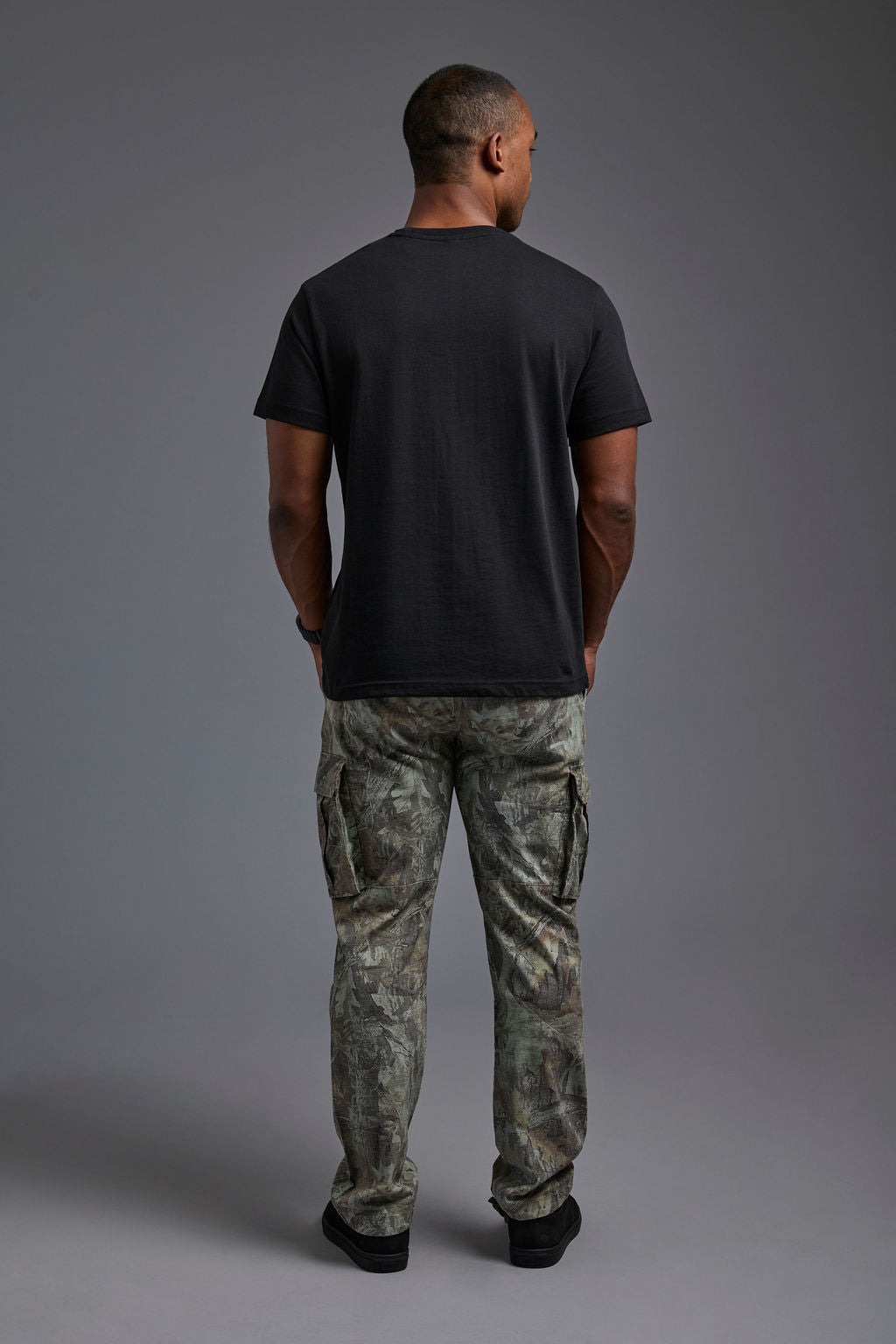 An African American male model with a beard wearing the men's core renewal tee in the color black and forest cargo pants from a back view on a gray background. The core renewal tee is made out of a sustainable blend of repurposed recycled garments: 60% recycled cotton and 40% recycled #black
