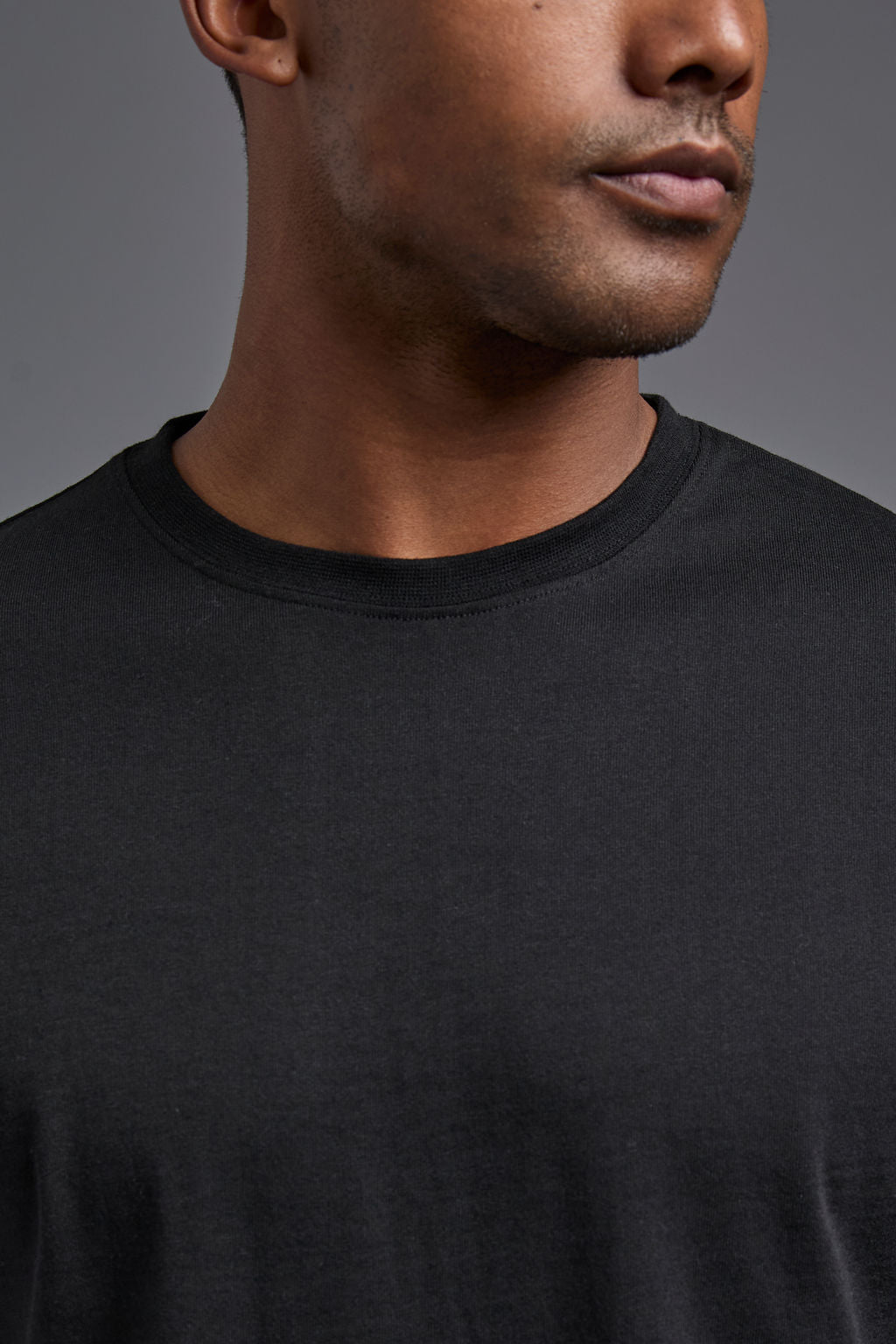 An African American male model with a beard wearing the men's core renewal tee in the color black and forest cargo pants from a front close up collar view on a gray background. The core renewal tee is made out of a sustainable blend of repurposed recycled garments: 60% recycled cotton and 40% recycled #black