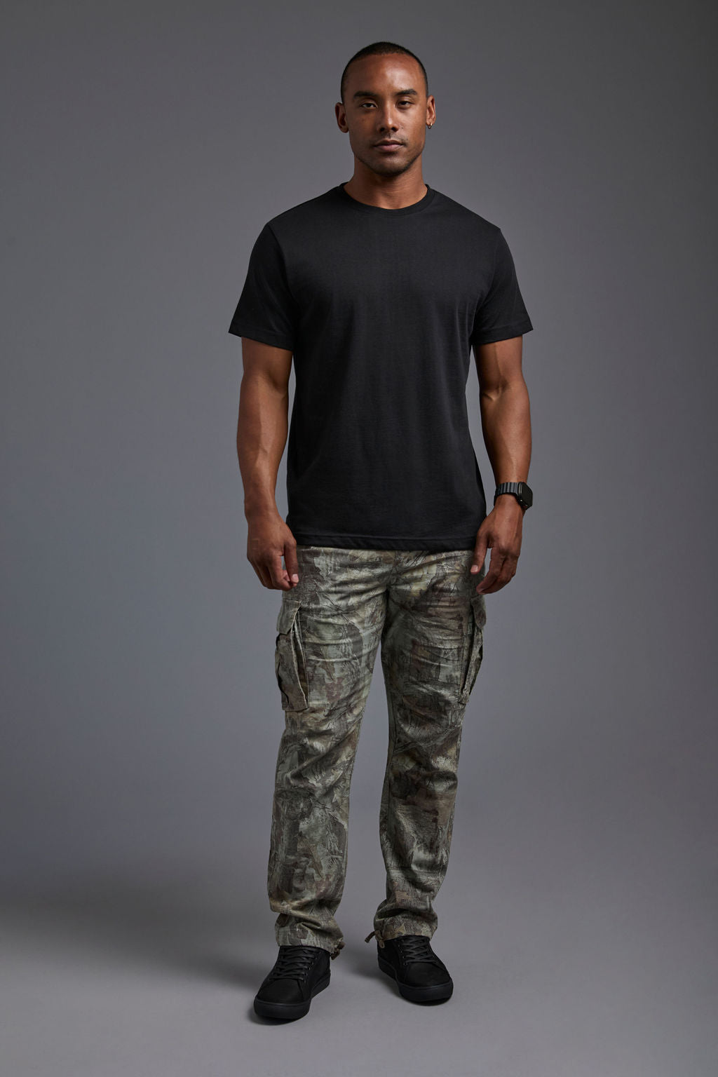 An African American male model with a beard wearing the men's core renewal tee in the color black and forest cargo pants from a front view on a gray background. The core renewal tee is made out of a sustainable blend of repurposed recycled garments: 60% recycled cotton and 40% recycled #black
