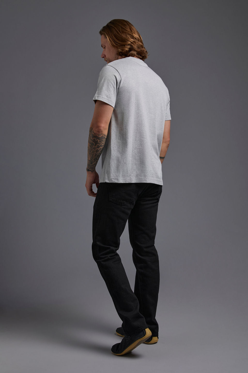 A blonde, male model with a beard wearing the men's core renewal tee in the color light grey melange heather and black Levi 501® jeans from a back view on a gray background. The core renewal tee is made out of a sustainable blend of repurposed recycled garments: 60% recycled cotton and 40% recycled polyester. #light-grey-melange