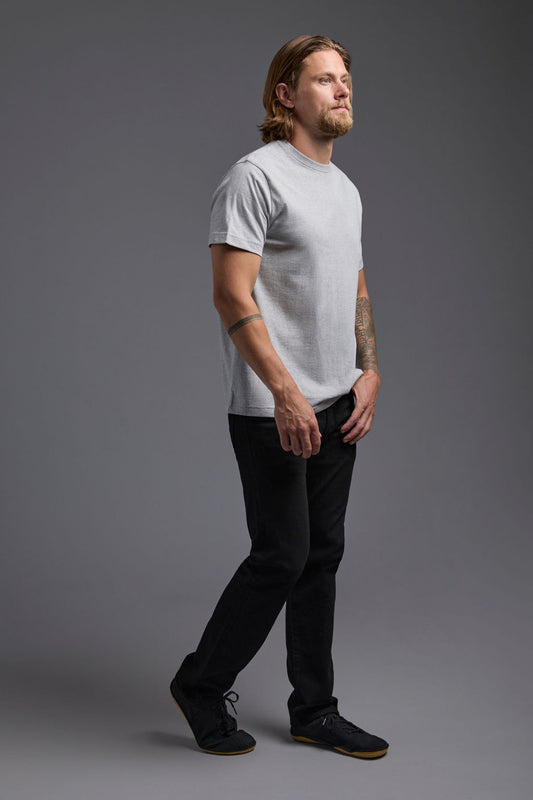 A blonde, male model with a beard wearing the men's core renewal tee in the color light grey melange heather and black Levi 501® jeans from a side view on a gray background. The core renewal tee is made out of a sustainable blend of repurposed recycled garments: 60% recycled cotton and 40% recycled polyester. #light-grey-melange