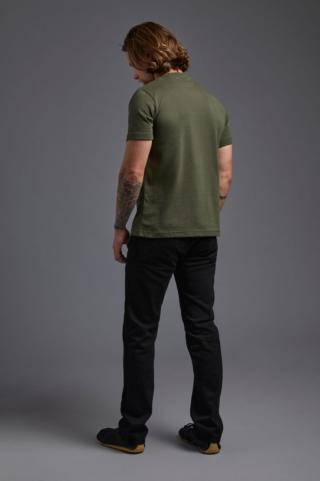 A blonde, male model with a beard wearing the men's core renewal tee in the color military green and black Levi 501® jeans from a back view on a gray background. The core renewal tee is made out of a sustainable blend of repurposed recycled garments: 60% recycled cotton and 40% recycled polyester. #military-green