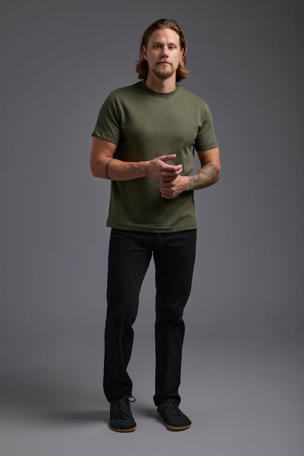 A blonde, male model with a beard wearing the men's core renewal tee in the color military green and black Levi 501® jeans from a front view on a gray background. The core renewal tee is made out of a sustainable blend of repurposed recycled garments: 60% recycled cotton and 40% recycled polyester. #military-green