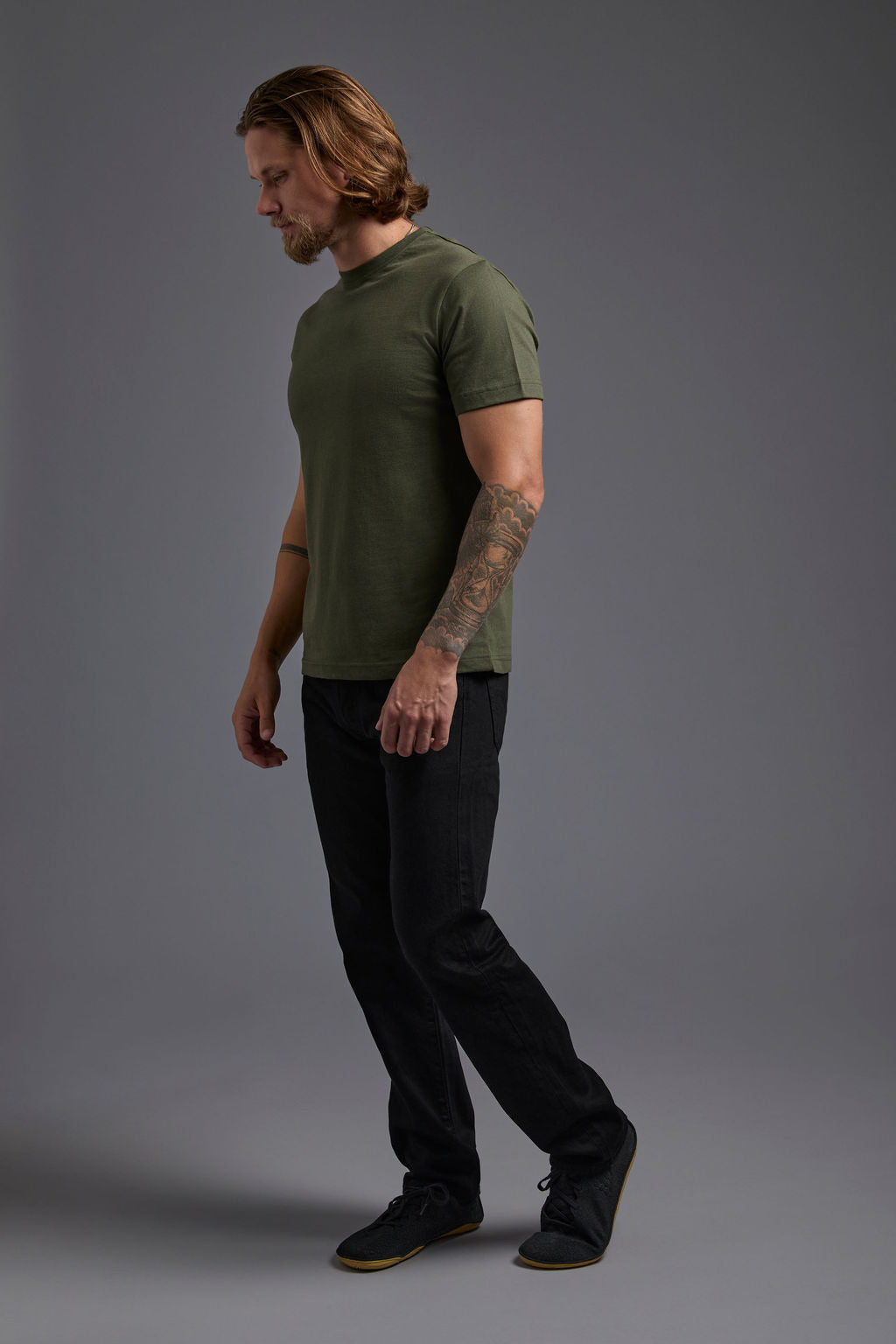 A blonde, male model with a beard wearing the men's core renewal tee in the color military green and black Levi 501® jeans from a side view on a gray background. The core renewal tee is made out of a sustainable blend of repurposed recycled garments: 60% recycled cotton and 40% recycled polyester. #military-green