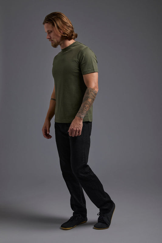 A blonde, male model with a beard wearing the men's core renewal tee in the color military green and black Levi 501® jeans from a side view on a gray background. The core renewal tee is made out of a sustainable blend of repurposed recycled garments: 60% recycled cotton and 40% recycled polyester. #military-green