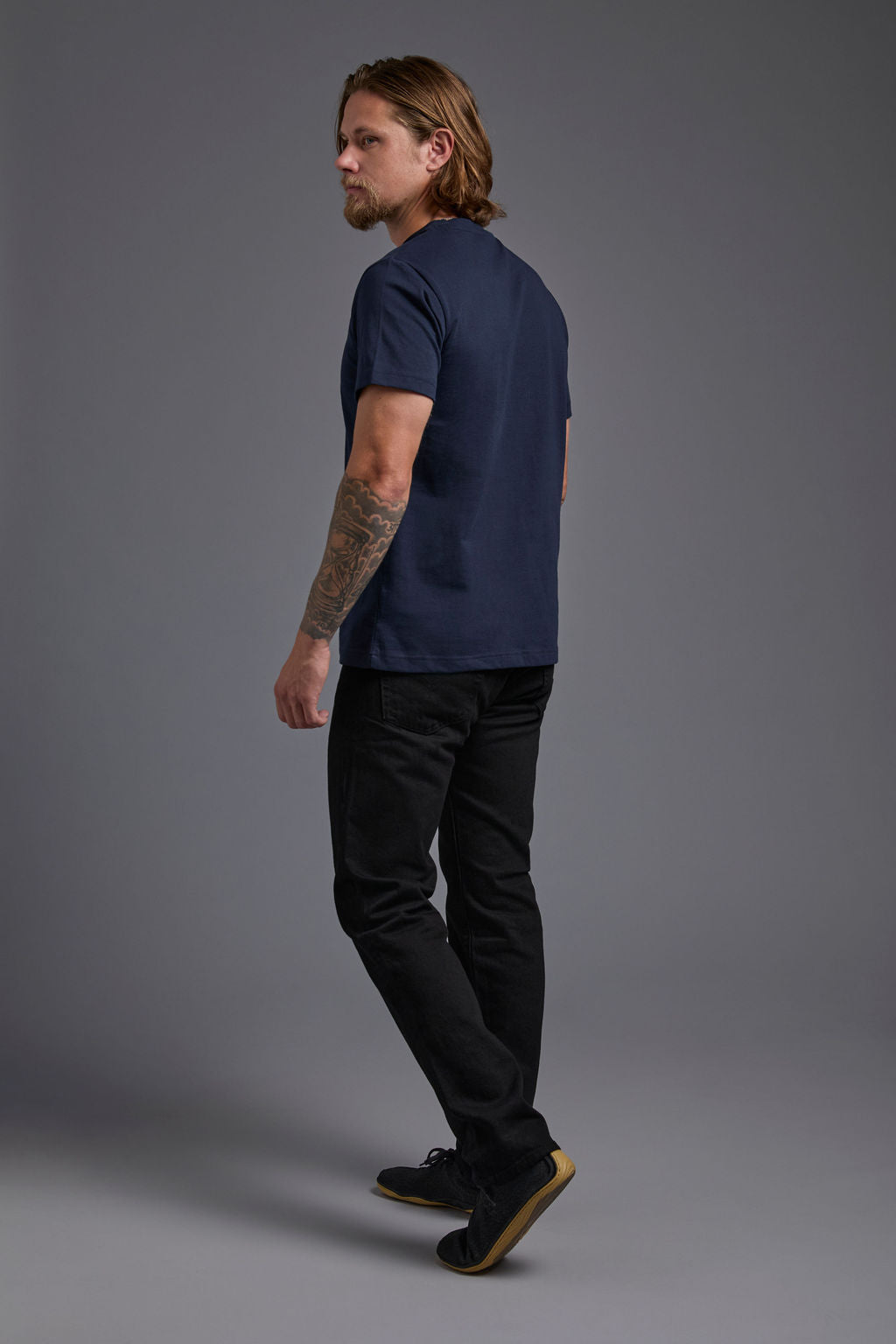 A blonde, male model with a beard wearing the men's core renewal tee in the color navy and black Levi 501® jeans from a back view on a gray background. The core renewal tee is made out of a sustainable blend of repurposed recycled garments: 60% recycled cotton and 40% recycled polyester. #navy