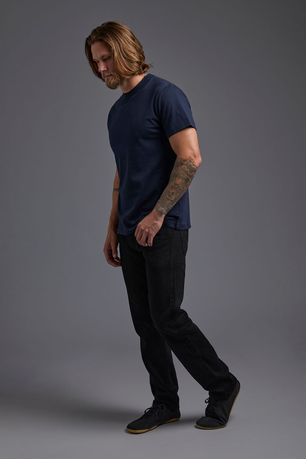 A blonde, male model with a beard wearing the men's core renewal tee in the color navy and black Levi 501® jeans from a side view on a gray background. The core renewal tee is made out of a sustainable blend of repurposed recycled garments: 60% recycled cotton and 40% recycled polyester. #navy