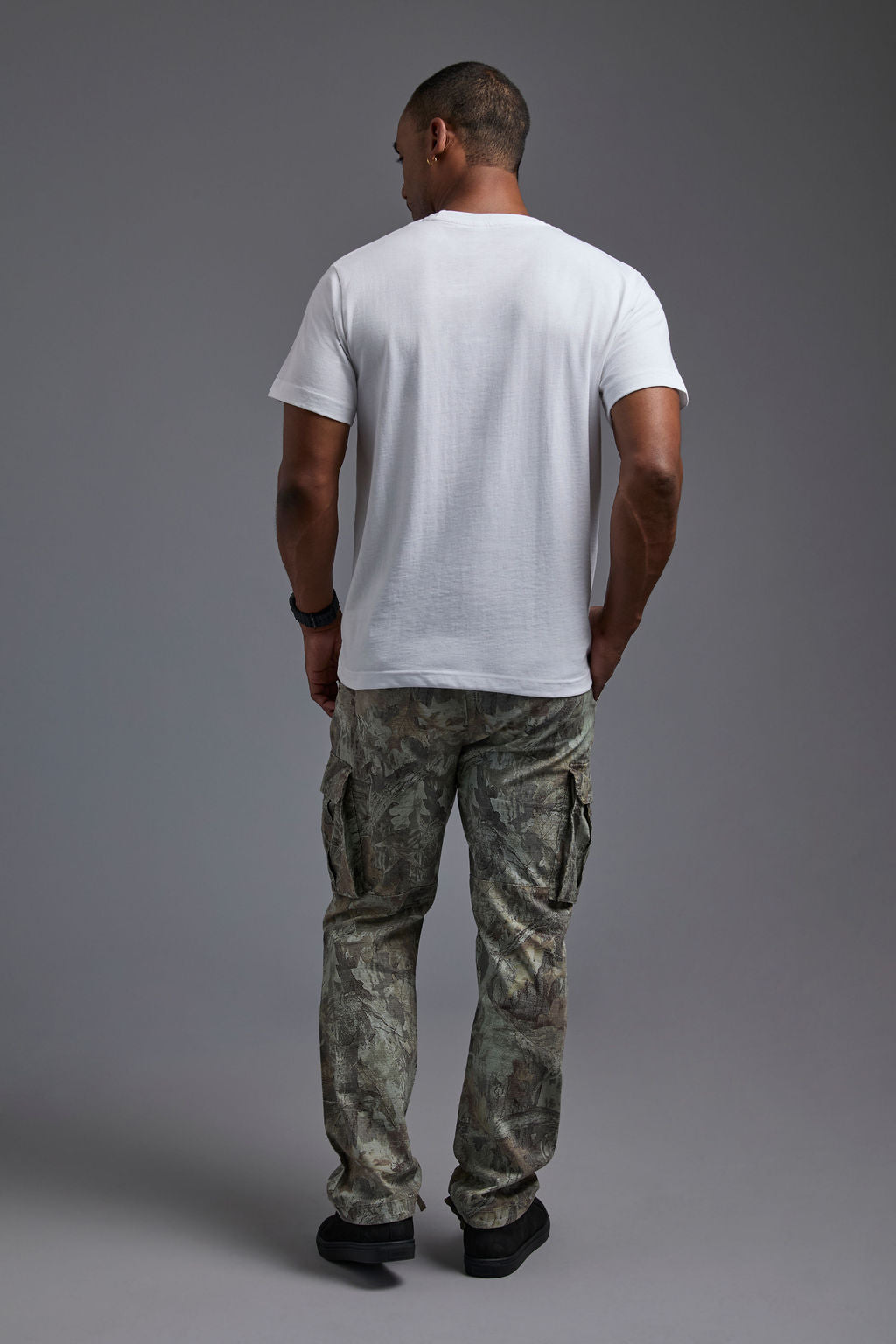 An African American male model with a beard wearing the men's core renewal tee in the color white and forest cargo pants from a back view on a gray background. The core renewal tee is made out of a sustainable blend of repurposed recycled garments: 60% recycled cotton and 40% recycled polyester. #white