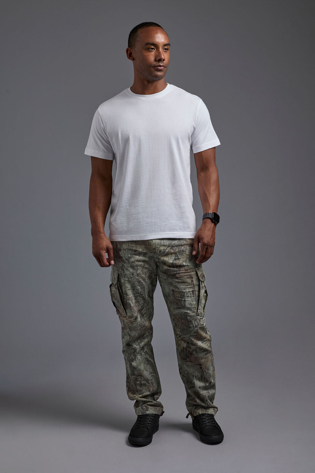 An African American male model with a beard wearing the men's core renewal tee in the color white and forest cargo pants from a front view on a gray background. The core renewal tee is made out of a sustainable blend of repurposed recycled garments: 60% recycled cotton and 40% recycled polyester. #white