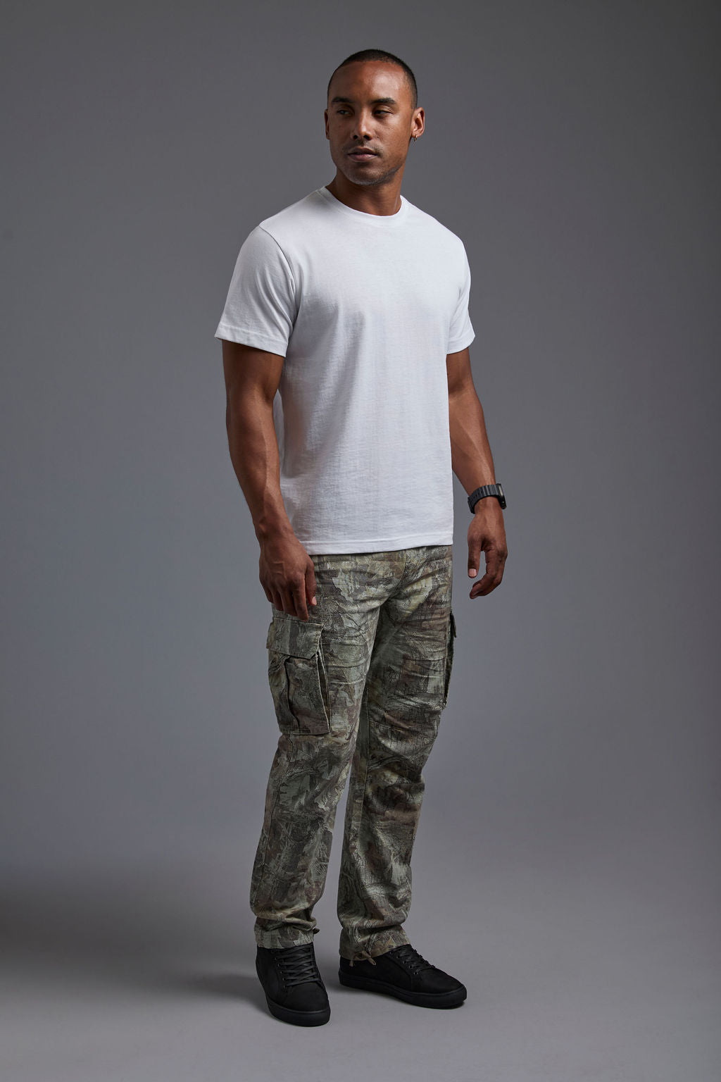 An African American male model with a beard wearing the men's core renewal tee in the color white and forest cargo pants from a side view on a gray background. The core renewal tee is made out of a sustainable blend of repurposed recycled garments: 60% recycled cotton and 40% recycled polyester. #white