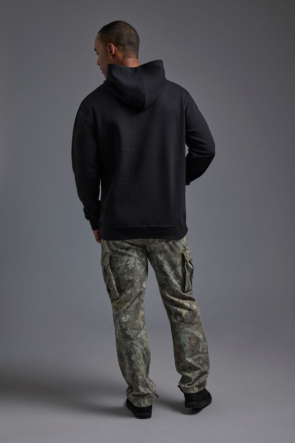 An African American, male model on a gray background wearing the men's renewal hoodie in the color black and forest cargo pants, made out of a sustainable blend of 60% recycled cotton and 40% recycled polyester, showing a relaxed fit from a back view with hood down. #black