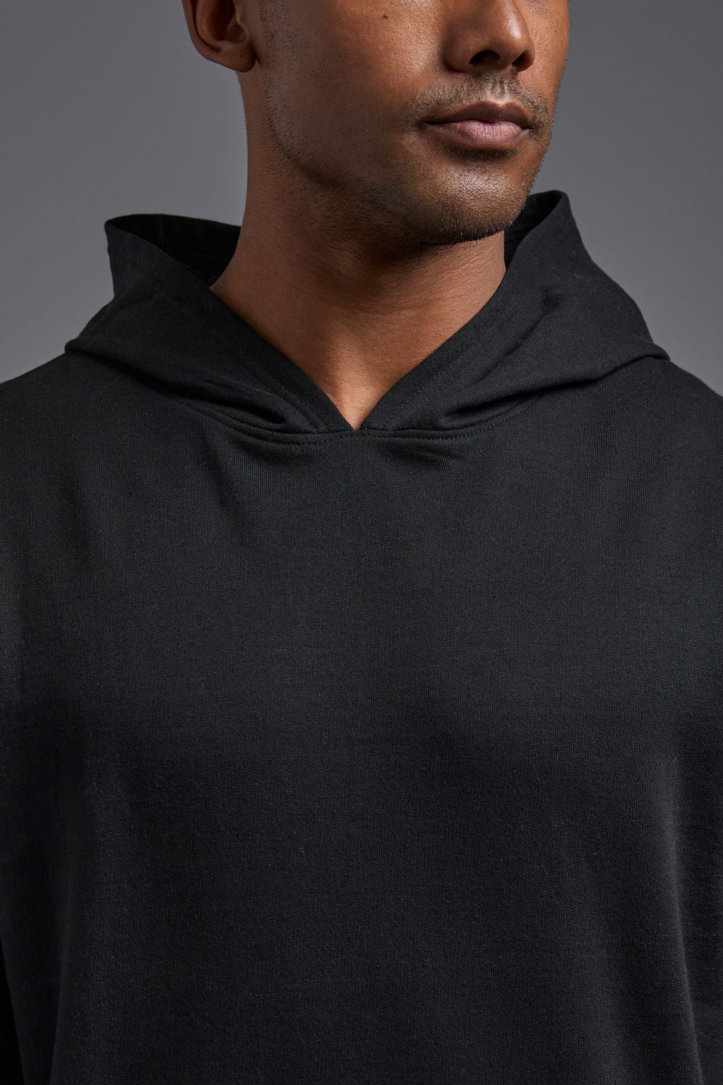 An African American, male model on a gray background wearing the men's renewal hoodie in the color black and forest cargo pants, made out of a sustainable blend of 60% recycled cotton and 40% recycled polyester, showing a relaxed fit from a front close up collar view. #black