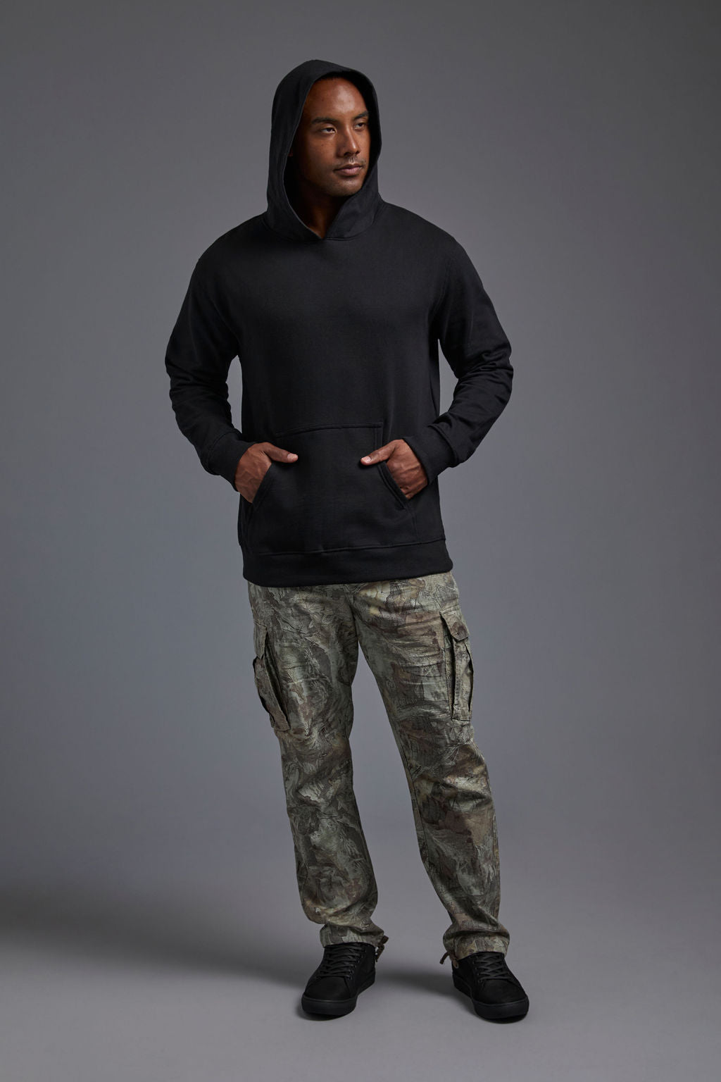 An African American, male model on a gray background wearing the men's renewal hoodie in the color black and forest cargo pants, made out of a sustainable blend of 60% recycled cotton and 40% recycled polyester, showing a relaxed fit from a front view with hood up. #black