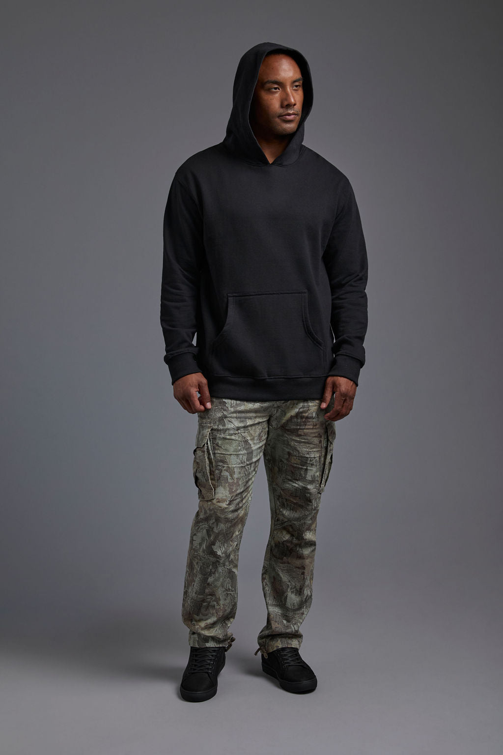 An African American, male model on a gray background wearing the men's renewal hoodie in the color black and forest cargo pants, made out of a sustainable blend of 60% recycled cotton and 40% recycled polyester, showing a relaxed fit from a front view with hood up and hands outside of kangaroo pocket. #black