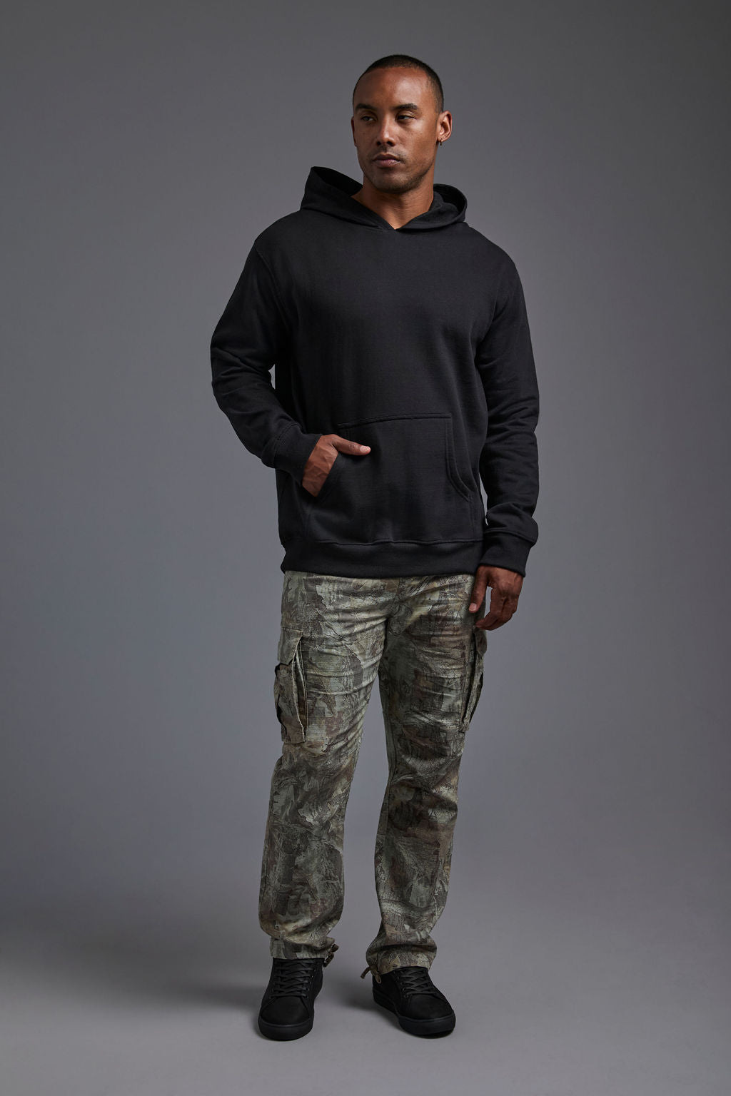 An African American, male model on a gray background wearing the men's renewal hoodie in the color black and forest cargo pants, made out of a sustainable blend of 60% recycled cotton and 40% recycled polyester, showing a relaxed fit from a front view. #black