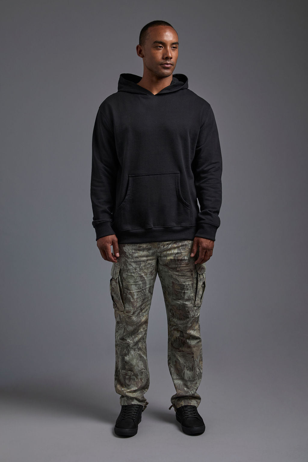 An African American, male model on a gray background wearing the men's renewal hoodie in the color black and forest cargo pants, made out of a sustainable blend of 60% recycled cotton and 40% recycled polyester, showing a relaxed fit from a front view with hood down. #black