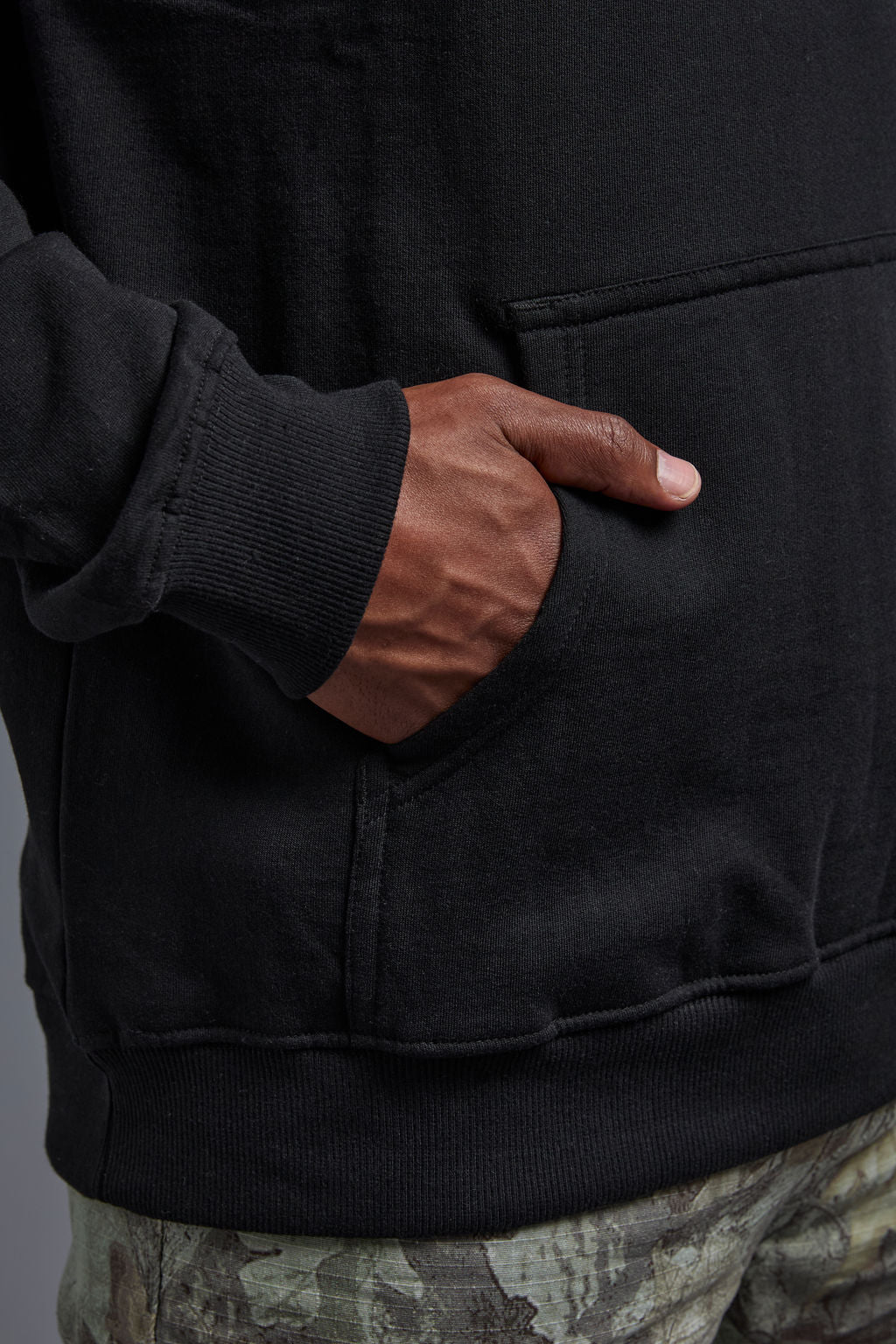 An African American, male model on a gray background wearing the men's renewal hoodie in the color black and forest cargo pants, made out of a sustainable blend of 60% recycled cotton and 40% recycled polyester, showing a relaxed fit from a front  kangaroo view. #black