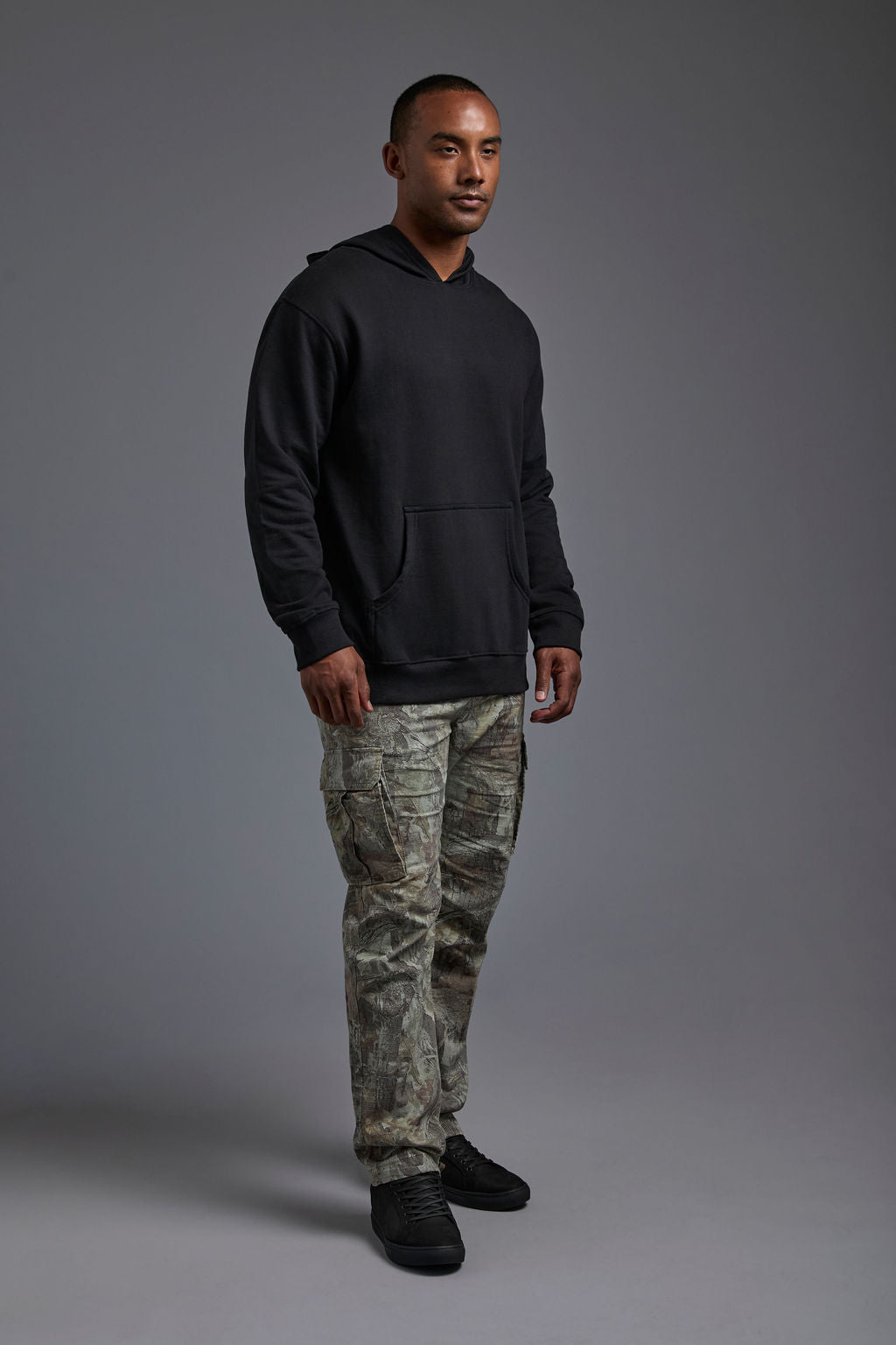 An African American, male model on a gray background wearing the men's renewal hoodie in the color black and forest cargo pants, made out of a sustainable blend of 60% recycled cotton and 40% recycled polyester, showing a relaxed fit from a side view. #black