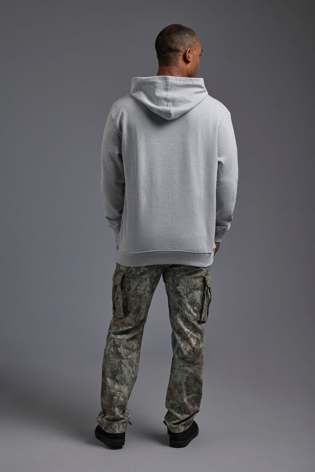An African American, male model on a gray background wearing the men's renewal hoodie in the color light grey melange and forest cargo pants, made out of a sustainable blend of 60% recycled cotton and 40% recycled polyester, showing a relaxed fit from a back view. #light-grey-melange