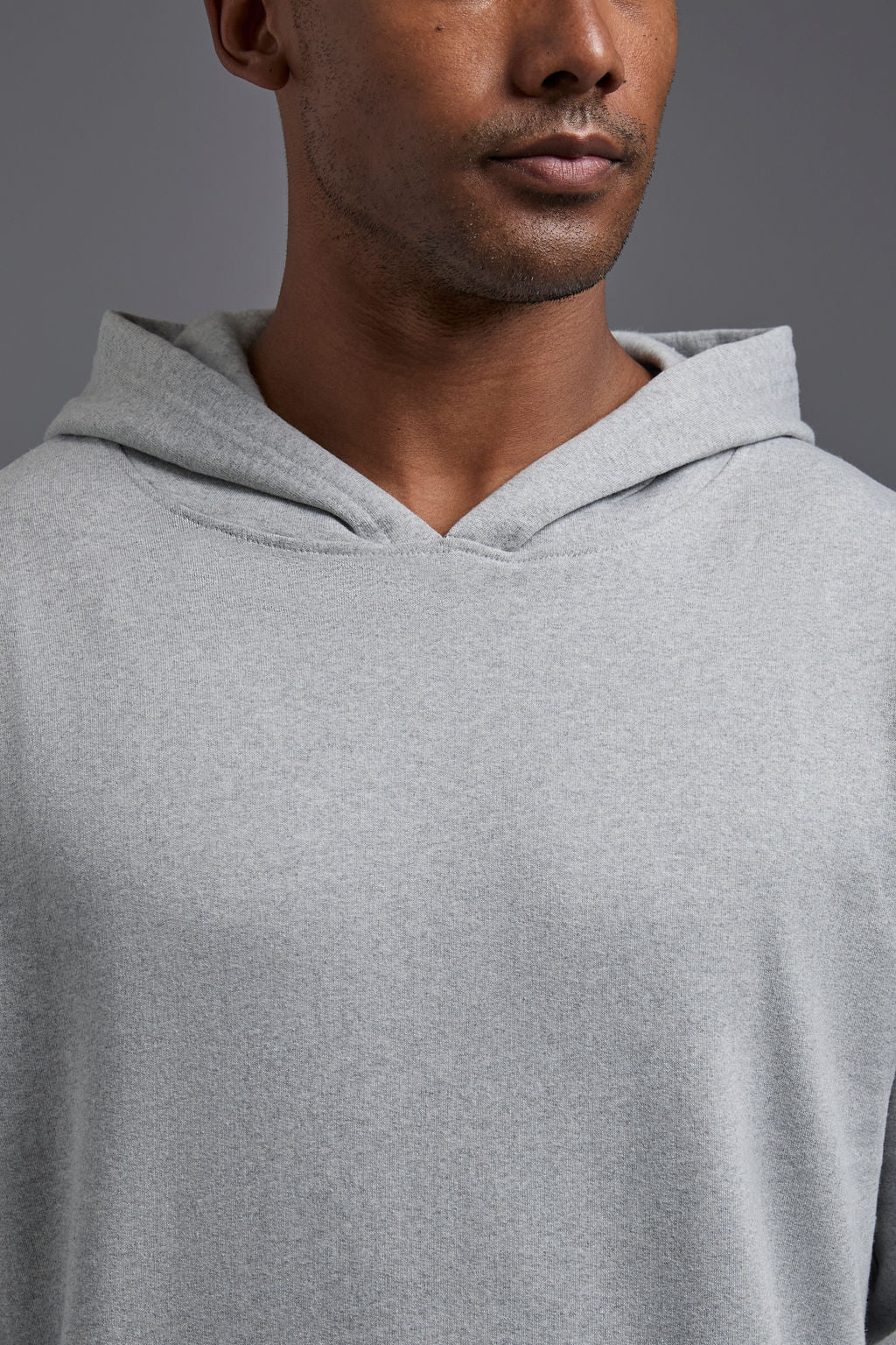 An African American, male model on a gray background wearing the men's renewal hoodie in the color light grey melange and forest cargo pants, made out of a sustainable blend of 60% recycled cotton and 40% recycled polyester, showing a relaxed fit from a front collar close up view. #light-grey-melange