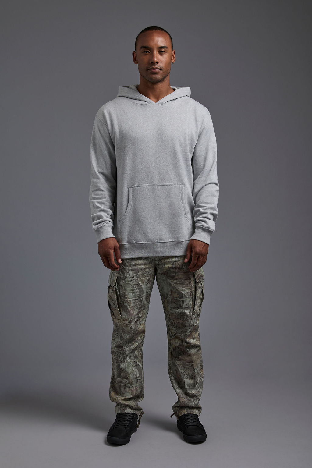 An African American, male model on a gray background wearing the men's renewal hoodie in the color light grey melange and forest cargo pants, made out of a sustainable blend of 60% recycled cotton and 40% recycled polyester, showing a relaxed fit from a front view. #light-grey-melange