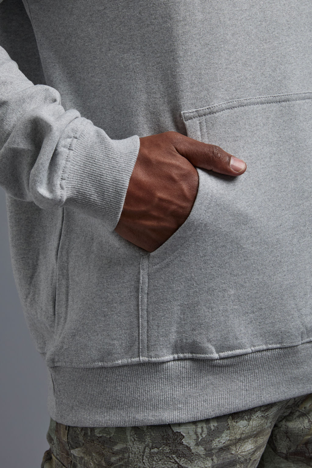 An African American, male model on a gray background wearing the men's renewal hoodie in the color light grey melange and forest cargo pants, made out of a sustainable blend of 60% recycled cotton and 40% recycled polyester, showing a relaxed fit from a front close up kangaroo pocket view. #light-grey-melange