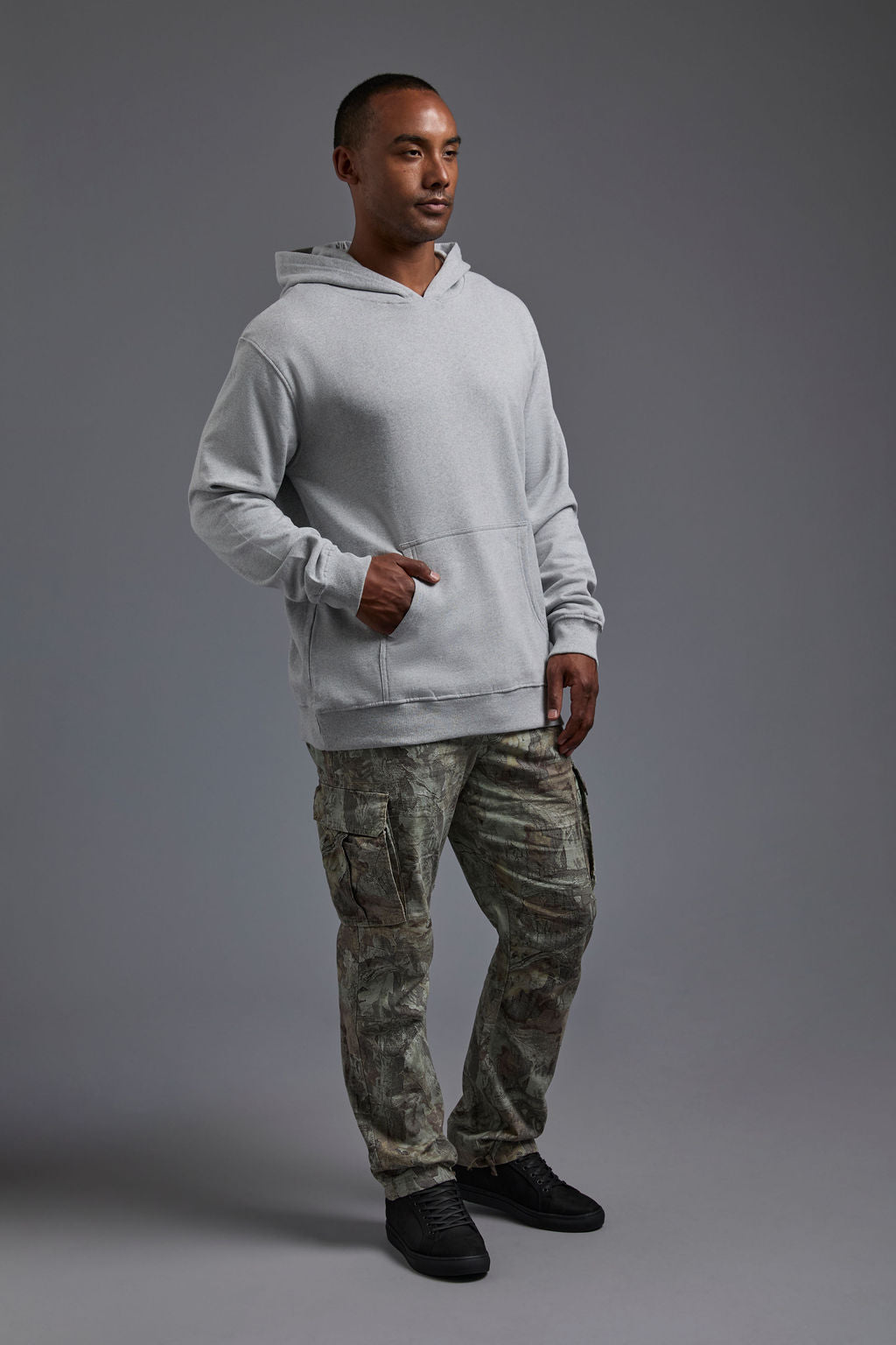 An African American, male model on a gray background wearing the men's renewal hoodie in the color light grey melange and forest cargo pants, made out of a sustainable blend of 60% recycled cotton and 40% recycled polyester, showing a relaxed fit from a side view. #light-grey-melange