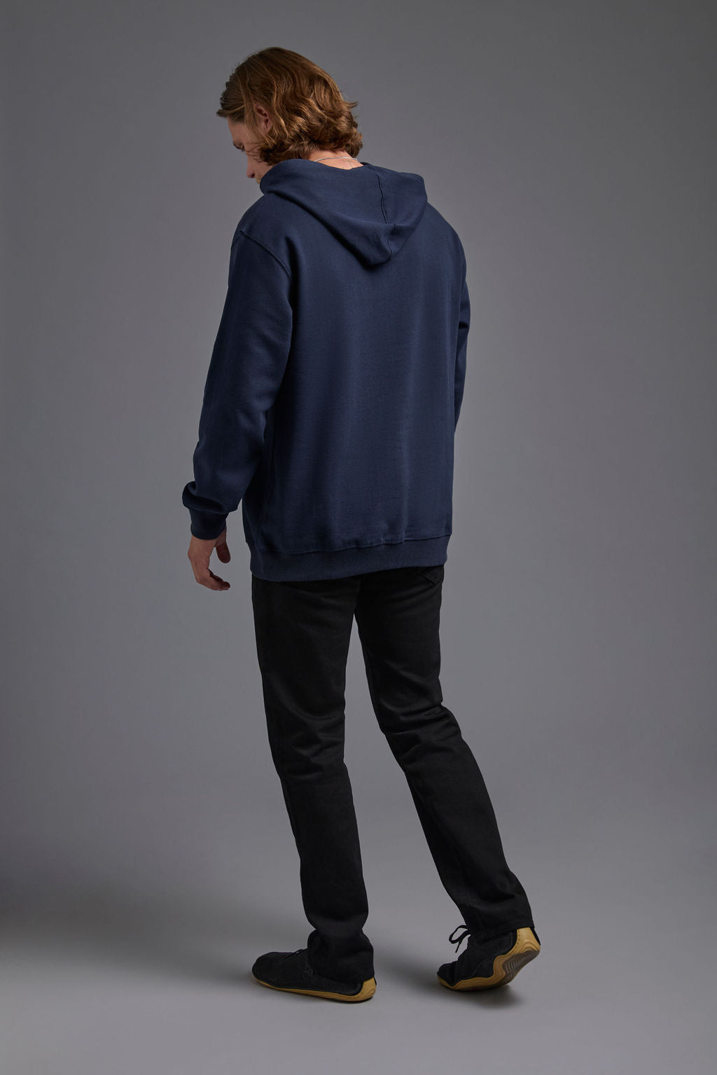 A blonde, male model with a beard on a gray background wearing the men's renewal hoodie in the color navy and black Levi 501® jeans. The renewal hoodie is made out of a sustainable blend of 60% recycled cotton and 40% recycled polyester, showing a back view. #navy