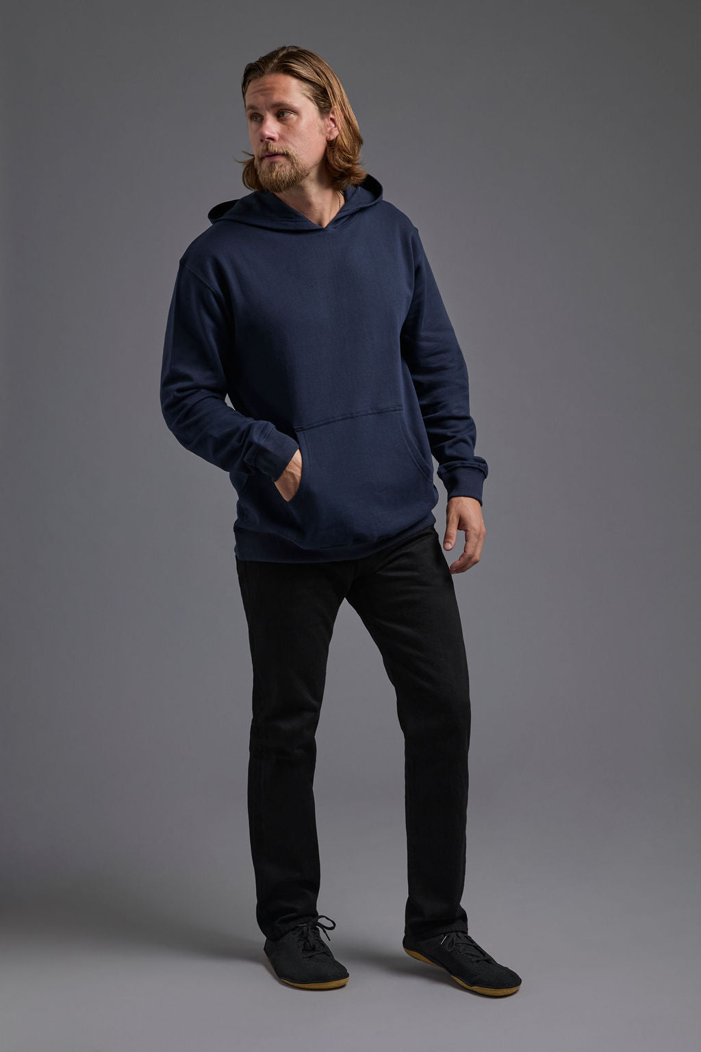 A blonde, male model with a beard on a gray background wearing the men's renewal hoodie in the color navy and black Levi 501® jeans. The renewal hoodie is made out of a sustainable blend of 60% recycled cotton and 40% recycled polyester, showing a relaxed fit from a side view. #navy