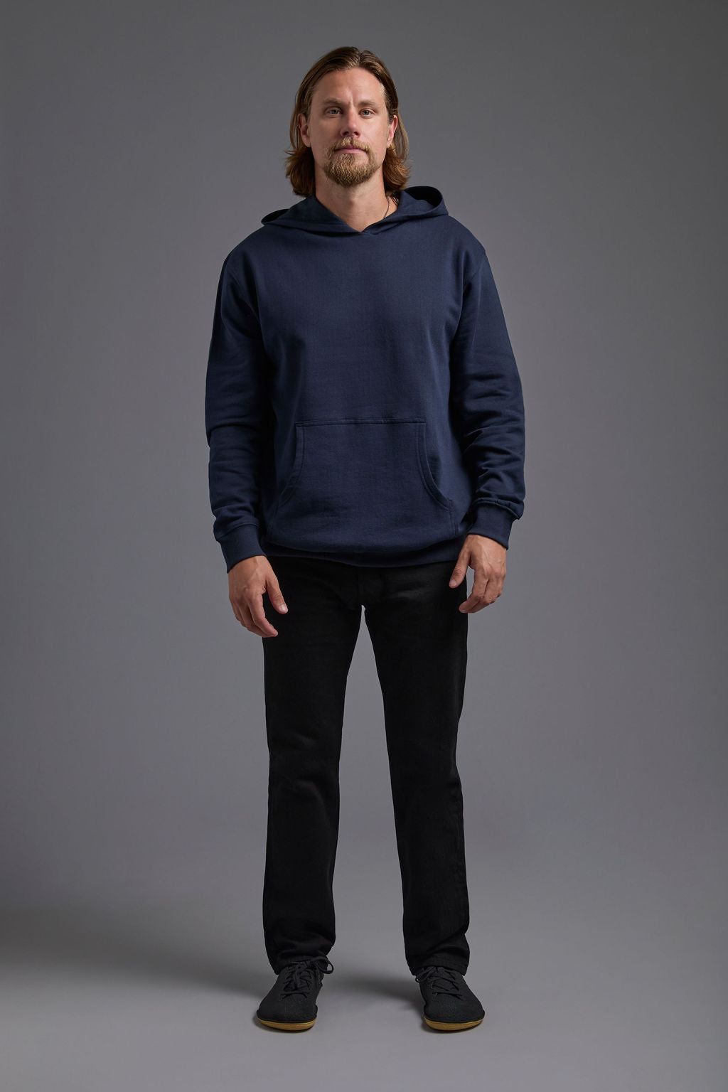 A blonde, male model with a beard on a gray background wearing the men's renewal hoodie in the color navy and black Levi 501® jeans. The renewal hoodie is made out of a sustainable blend of 60% recycled cotton and 40% recycled polyester, showing a relaxed fit from a front view. #navy