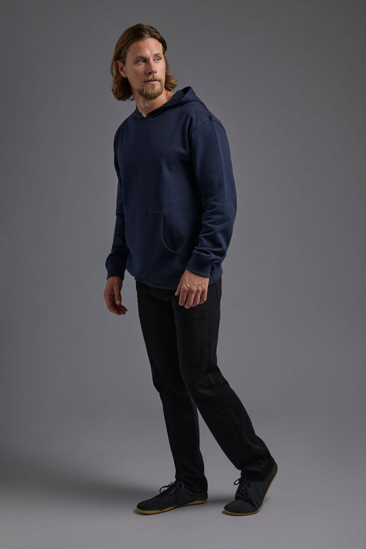 A blonde, male model with a beard on a gray background wearing the men's renewal hoodie in the color navy and black Levi 501® jeans. The renewal hoodie is made out of a sustainable blend of 60% recycled cotton and 40% recycled polyester, showing a relaxed fit from a side view. #navy