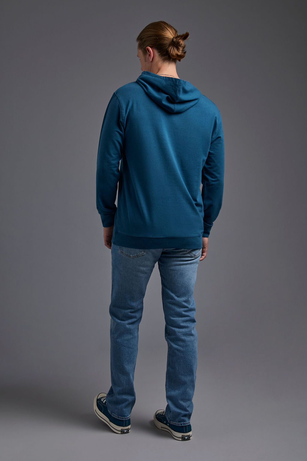 A blonde, male model with a beard on a gray background wearing the men's premium bamboo stretch hoodie in the color atlantic blue, made out of 45% Bamboo, 50% Cotton, 5% Spandex, showing a relaxed fit from a back view. The model is wearing levi's 501 jeans and teal converse high top sneakers. #atlantic-blue