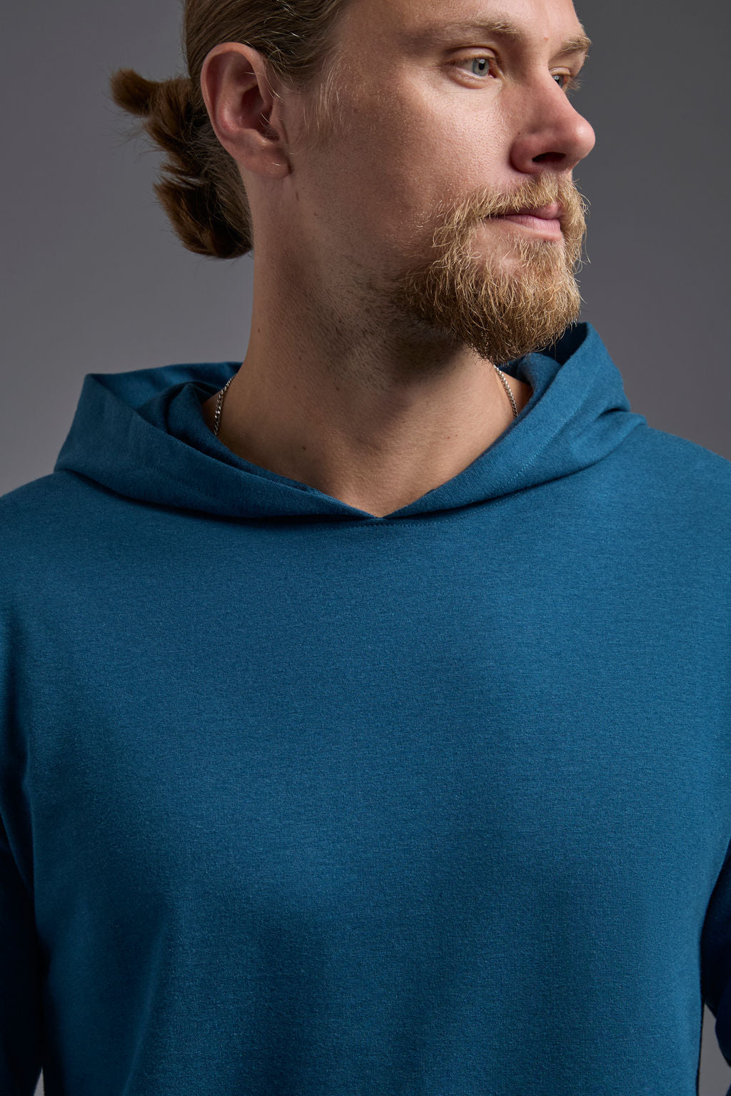 A blonde, male model with a beard on a gray background wearing the men's premium bamboo stretch hoodie in the color atlantic blue, made out of 45% Bamboo, 50% Cotton, 5% Spandex, showing a relaxed fit from a close up collar front view. The model is wearing levi's 501 jeans and teal converse high top sneakers. #atlantic-blue