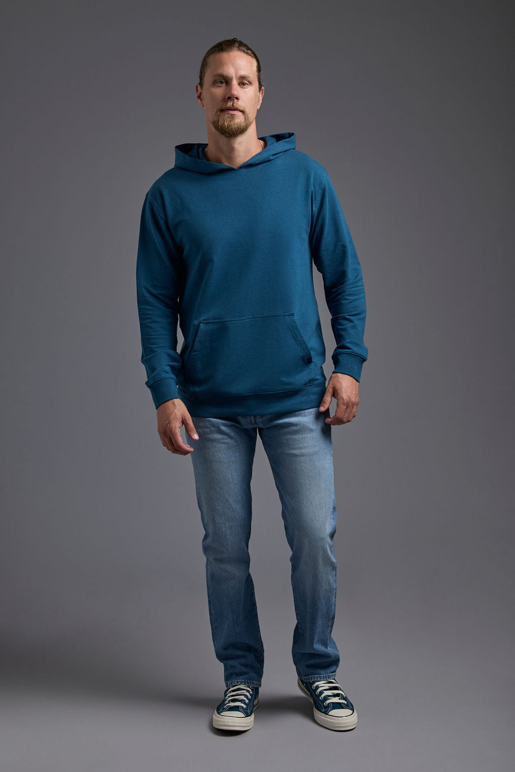 A blonde, male model with a beard on a gray background wearing the men's premium bamboo stretch hoodie in the color atlantic blue, made out of 45% Bamboo, 50% Cotton, 5% Spandex, showing a relaxed fit from a front view. The model is wearing levi's 501 jeans and teal converse high top sneakers. #atlantic-blue
