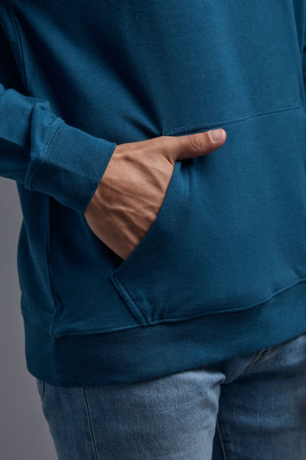A blonde, male model with a beard on a gray background wearing the men's premium bamboo stretch hoodie in the color atlantic blue, made out of 45% Bamboo, 50% Cotton, 5% Spandex, showing a relaxed fit from a front close up pocket view. The model is wearing levi's 501 jeans and teal converse high top sneakers. #atlantic-blue