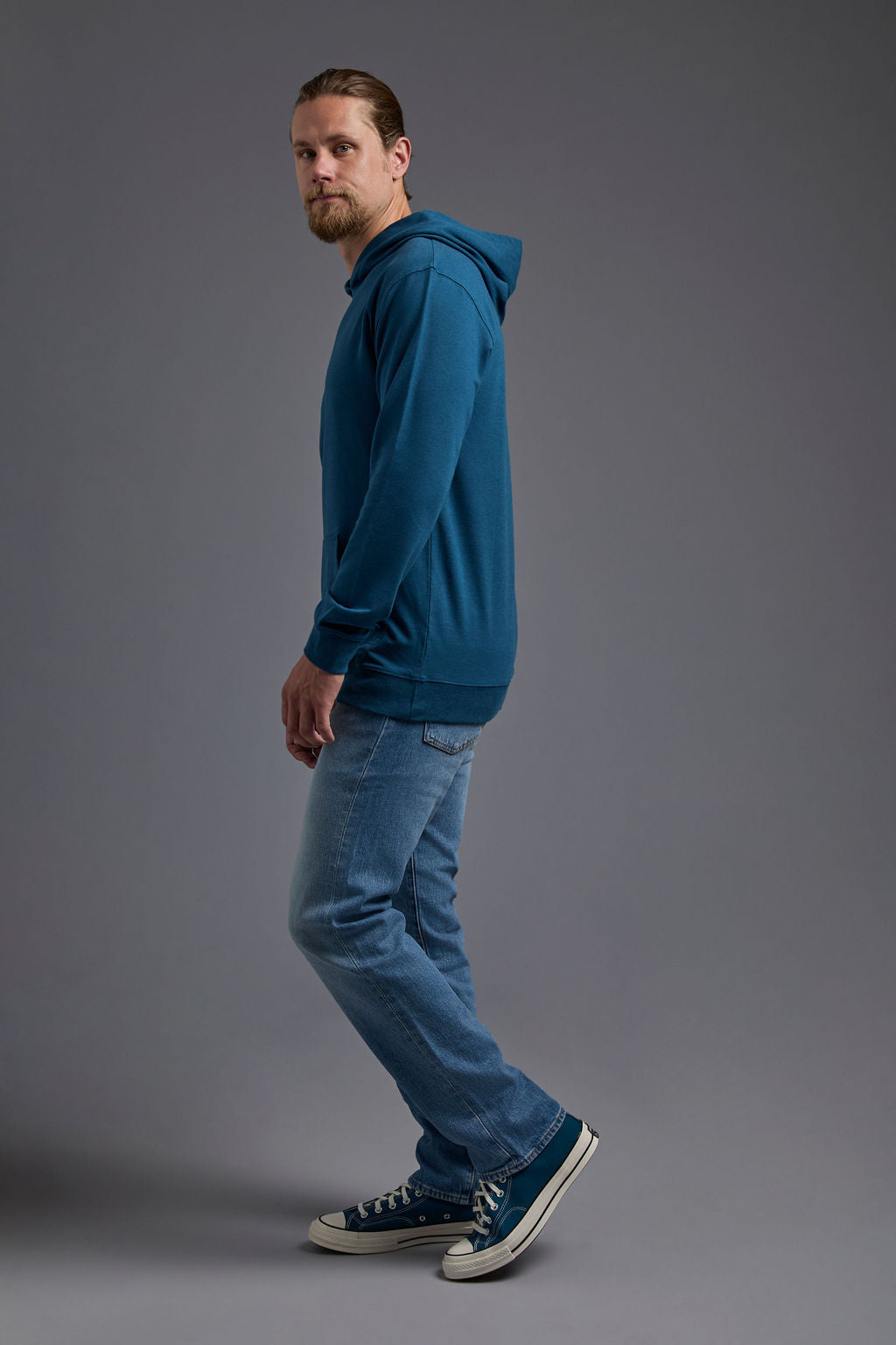 A blonde, male model with a beard on a gray background wearing the men's premium bamboo stretch hoodie in the color atlantic blue, made out of 45% Bamboo, 50% Cotton, 5% Spandex, showing a relaxed fit from a side view. The model is wearing levi's 501 jeans and teal converse high top sneakers. #atlantic-blue