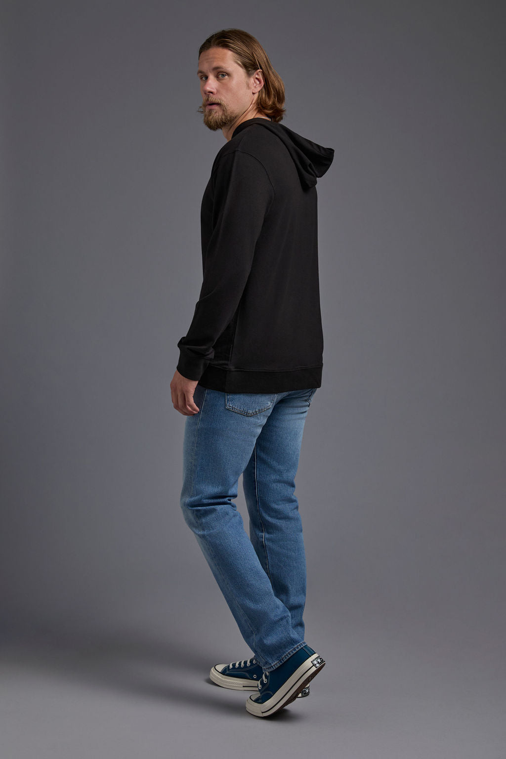 A blonde, male model with a beard on a gray background wearing the men's premium bamboo stretch hoodie in the color black, made out of 45% Bamboo, 50% Cotton, 5% Spandex, showing a relaxed fit from a side view. The model is wearing levi's 501 jeans and teal converse high top sneakers. #black
