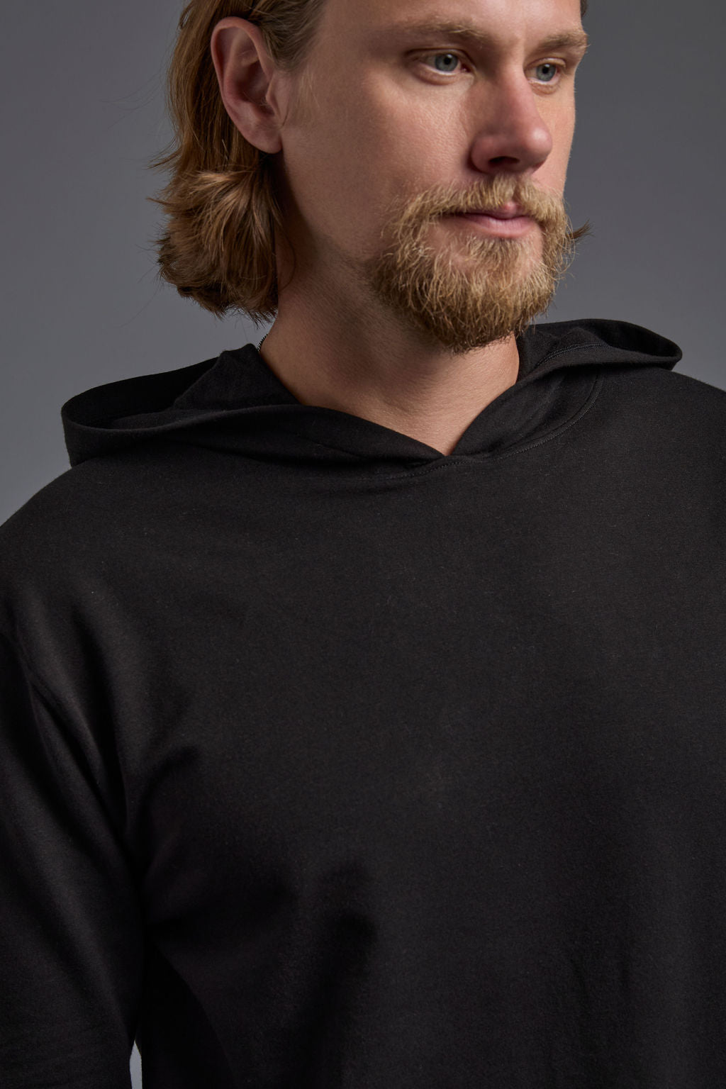 A blonde, male model with a beard on a gray background wearing the men's premium bamboo stretch hoodie in the color black, made out of 45% Bamboo, 50% Cotton, 5% Spandex, showing a relaxed fit from a front close up collar view. The model is wearing levi's 501 jeans and teal converse high top sneakers. #black