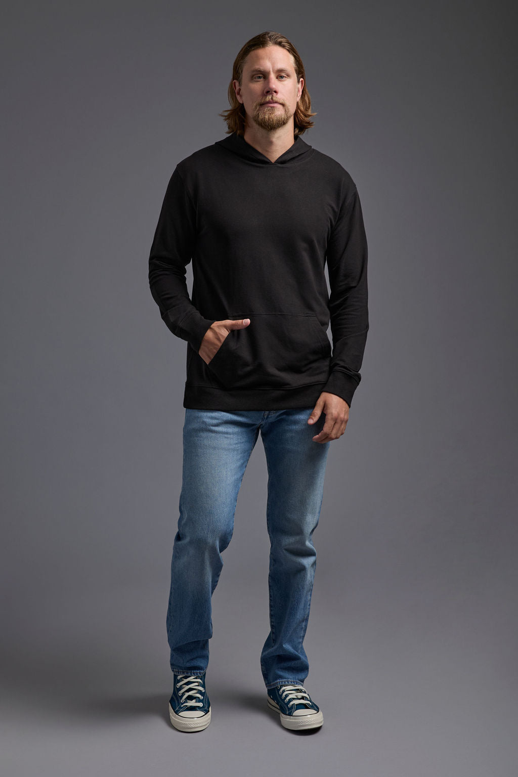 A blonde, male model with a beard on a gray background wearing the men's premium bamboo stretch hoodie in the color black, made out of 45% Bamboo, 50% Cotton, 5% Spandex, showing a relaxed fit from a front view. The model is wearing levi's 501 jeans and teal converse high top sneakers. #black
