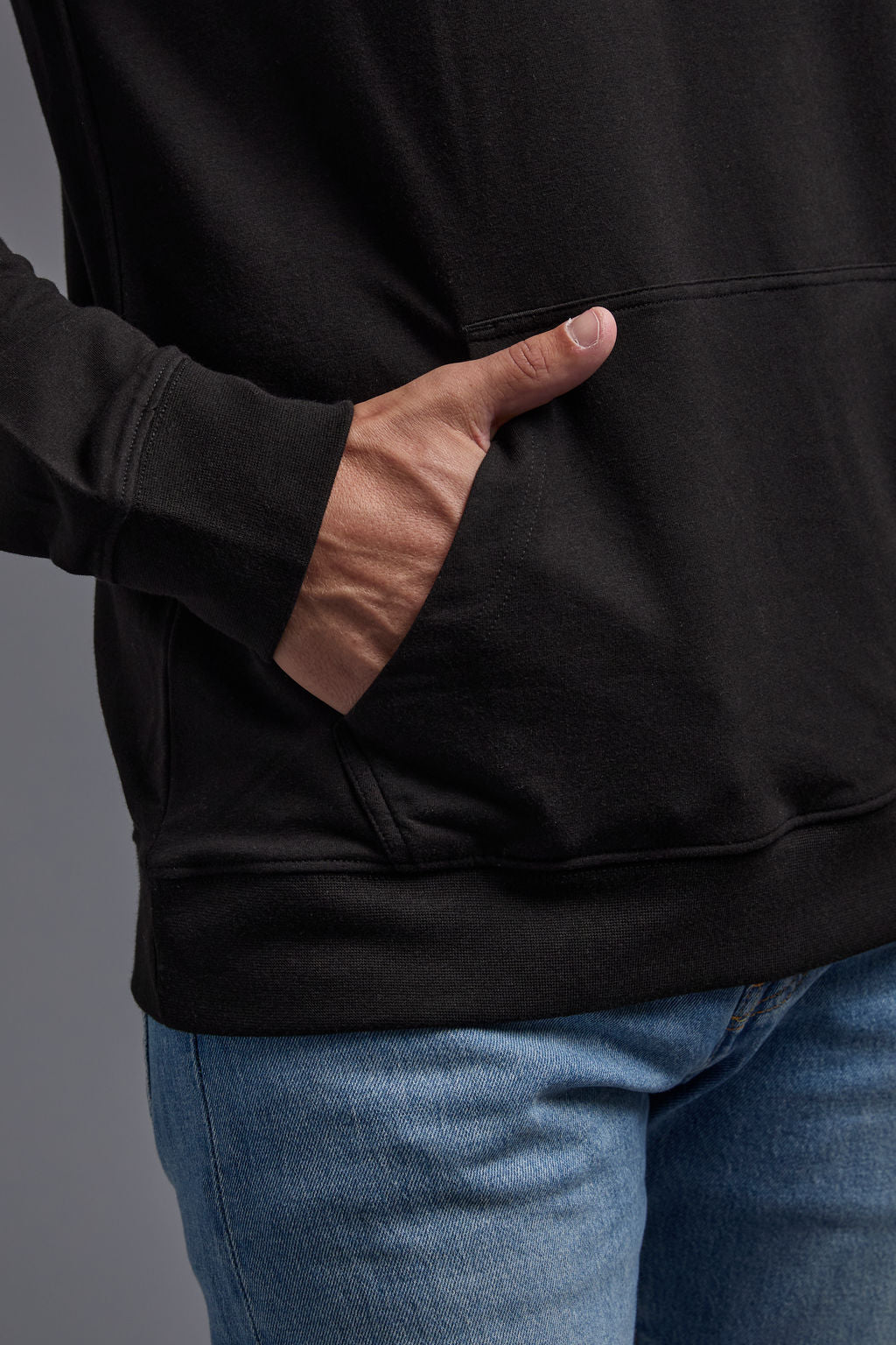 A blonde, male model with a beard on a gray background wearing the men's premium bamboo stretch hoodie in the color black, made out of 45% Bamboo, 50% Cotton, 5% Spandex, showing a relaxed fit from a front kangaroo pocket view. The model is wearing levi's 501 jeans and teal converse high top sneakers. #black