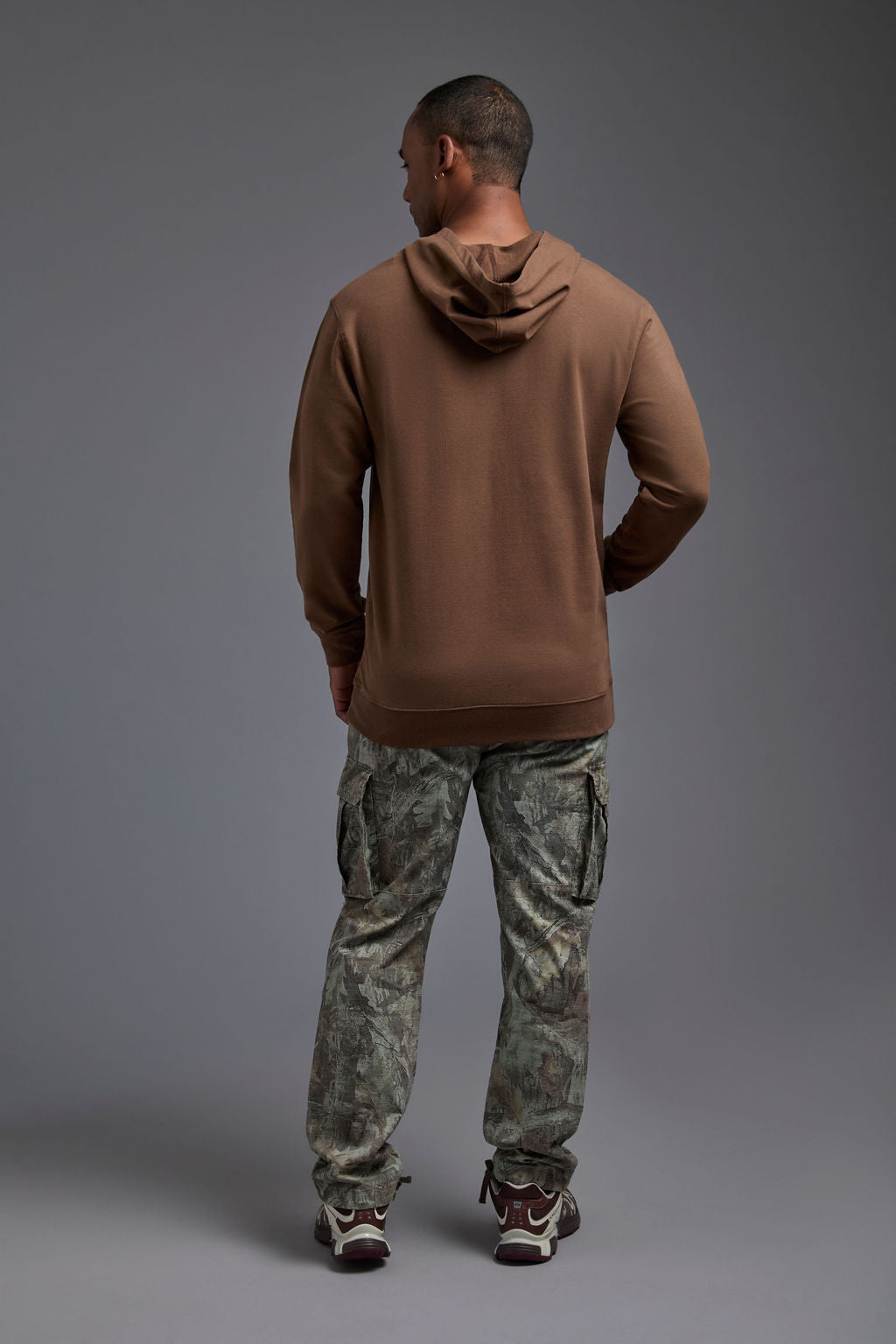 An African American male model on a gray background wearing the men's premium bamboo stretch hoodie in the color mocha brown, made out of 45% Bamboo, 50% Cotton, 5% Spandex, showing a relaxed fit from a back view. The model is wearing forest cargo pants and adidas sneakers. #mocha