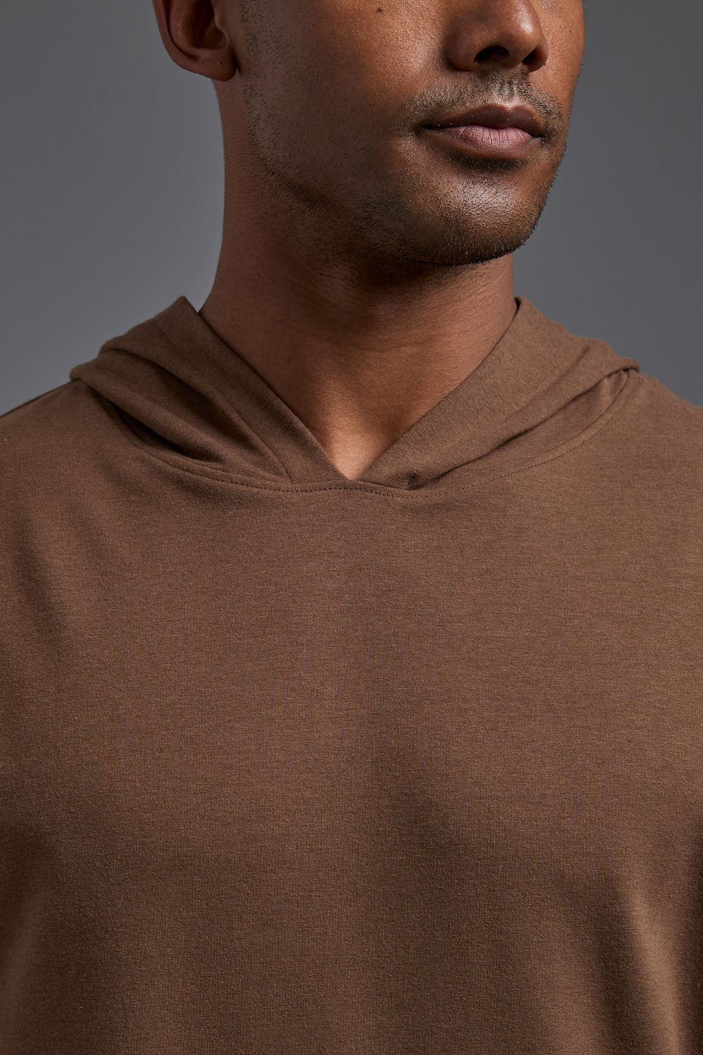 An African American male model on a gray background wearing the men's premium bamboo stretch hoodie in the color mocha brown, made out of 45% Bamboo, 50% Cotton, 5% Spandex, showing a relaxed fit from a front collar close up view. The model is wearing forest cargo pants and adidas sneakers. #mocha