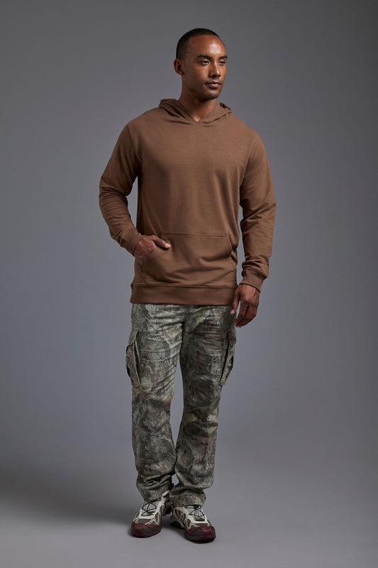 An African American male model on a gray background wearing the men's premium bamboo stretch hoodie in the color mocha brown, made out of 45% Bamboo, 50% Cotton, 5% Spandex, showing a relaxed fit from a front view. The model is wearing forest cargo pants and adidas sneakers. #mocha