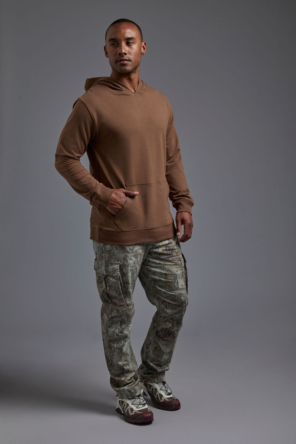 An African American male model on a gray background wearing the men's premium bamboo stretch hoodie in the color mocha brown, made out of 45% Bamboo, 50% Cotton, 5% Spandex, showing a relaxed fit from a side view. The model is wearing forest cargo pants and adidas sneakers. #mocha