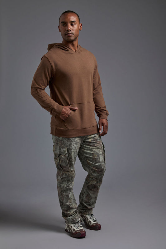 An African American male model on a gray background wearing the men's premium bamboo stretch hoodie in the color mocha brown, made out of 45% Bamboo, 50% Cotton, 5% Spandex, showing a relaxed fit from a side view. The model is wearing forest cargo pants and adidas sneakers. #mocha