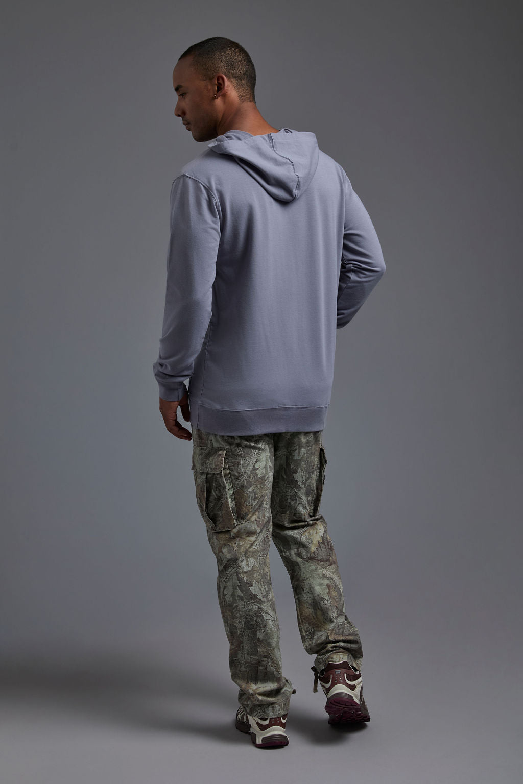 An African American male model on a gray background wearing the men's premium bamboo stretch hoodie in the color slate blue, made out of 45% Bamboo, 50% Cotton, 5% Spandex, showing a relaxed fit from a back view. The model is wearing forest cargo pants and adidas sneakers. #slate-blue