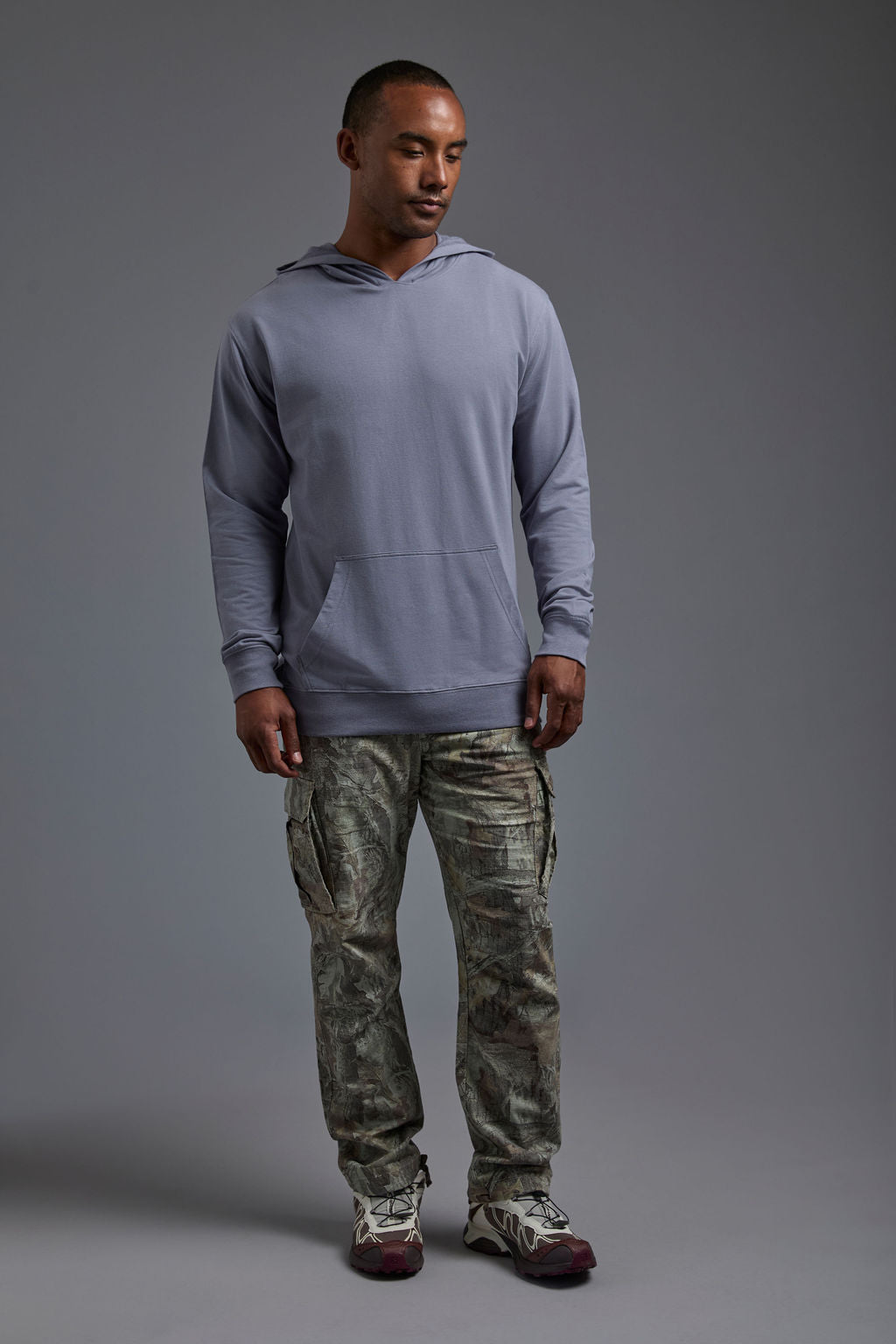 An African American male model on a gray background wearing the men's premium bamboo stretch hoodie in the color slate blue, made out of 45% Bamboo, 50% Cotton, 5% Spandex, showing a relaxed fit from a front view. The model is wearing forest cargo pants and adidas sneakers. #slate-blue