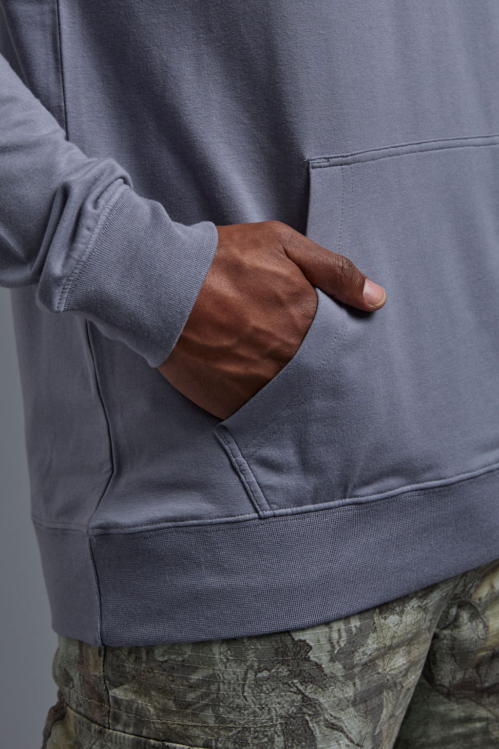 An African American male model on a gray background wearing the men's premium bamboo stretch hoodie in the color slate blue, made out of 45% Bamboo, 50% Cotton, 5% Spandex, showing a relaxed fit from a front kangaroo pocket close up view. The model is wearing forest cargo pants and adidas sneakers. #slate-blue