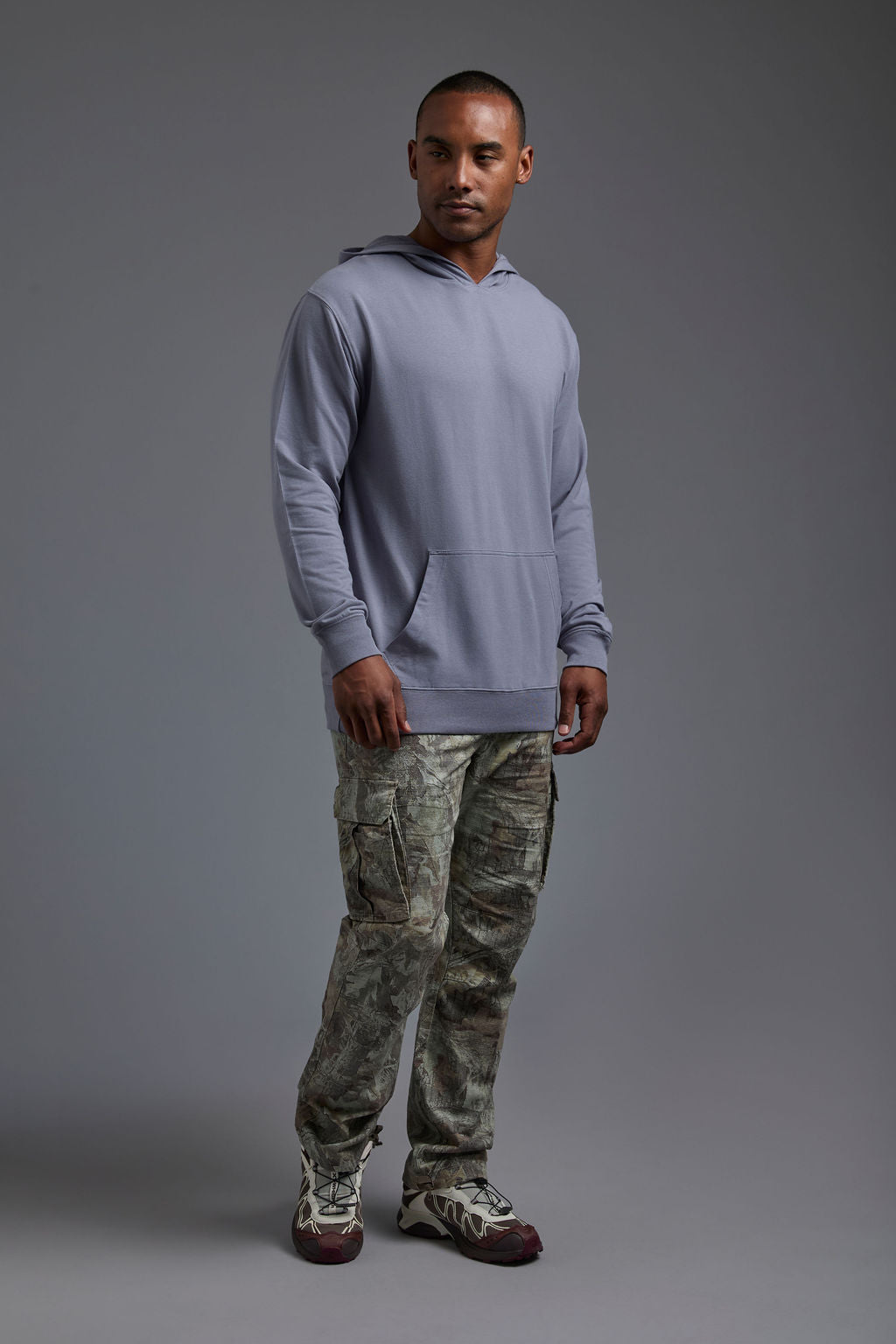 An African American male model on a gray background wearing the men's premium bamboo stretch hoodie in the color slate blue, made out of 45% Bamboo, 50% Cotton, 5% Spandex, showing a relaxed fit from a side view. The model is wearing forest cargo pants and adidas sneakers. #slate-blue