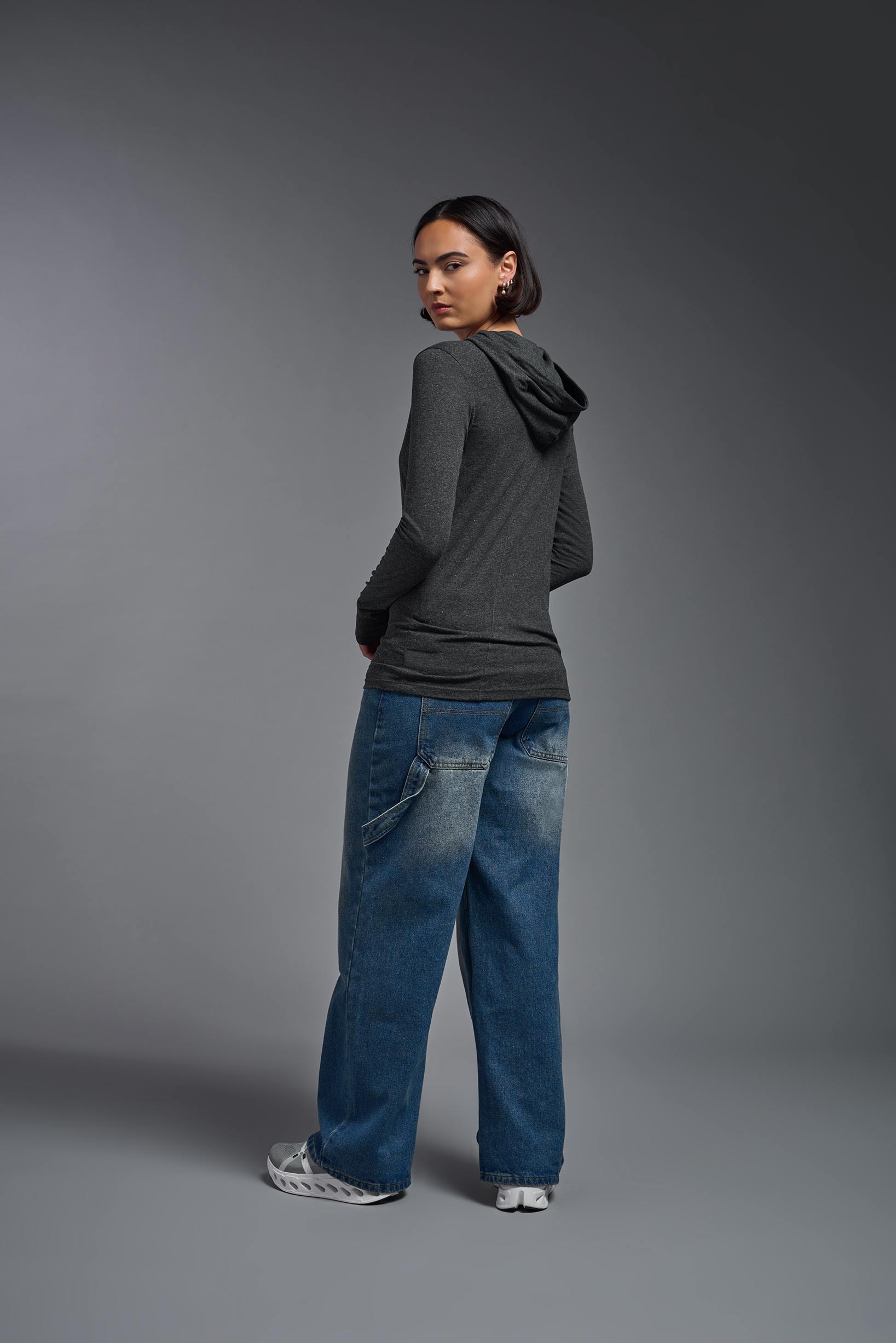 A female model wearing the women's Heather Hoodie Shirt in the color dark heather charcoal, made out of 88% polyester and 22% treated cotton, showing the relaxed fit from a back view. This hoodie shirt is comfortable and made in the USA. #dark-heather-charcoal